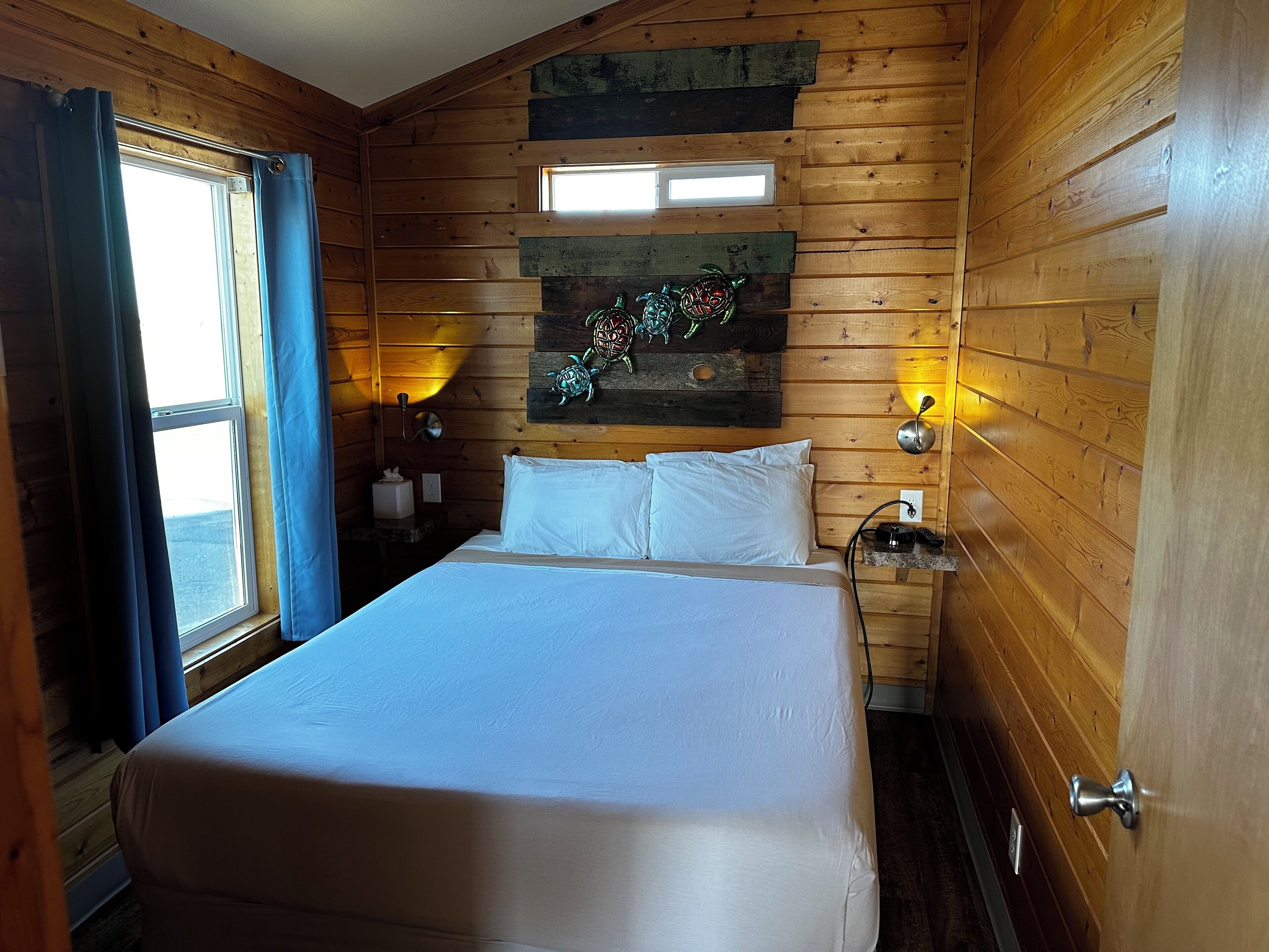 A wooden-paneled room with a bed, a window with curtains, a wall-mounted light, and a decorative piece above the bed.