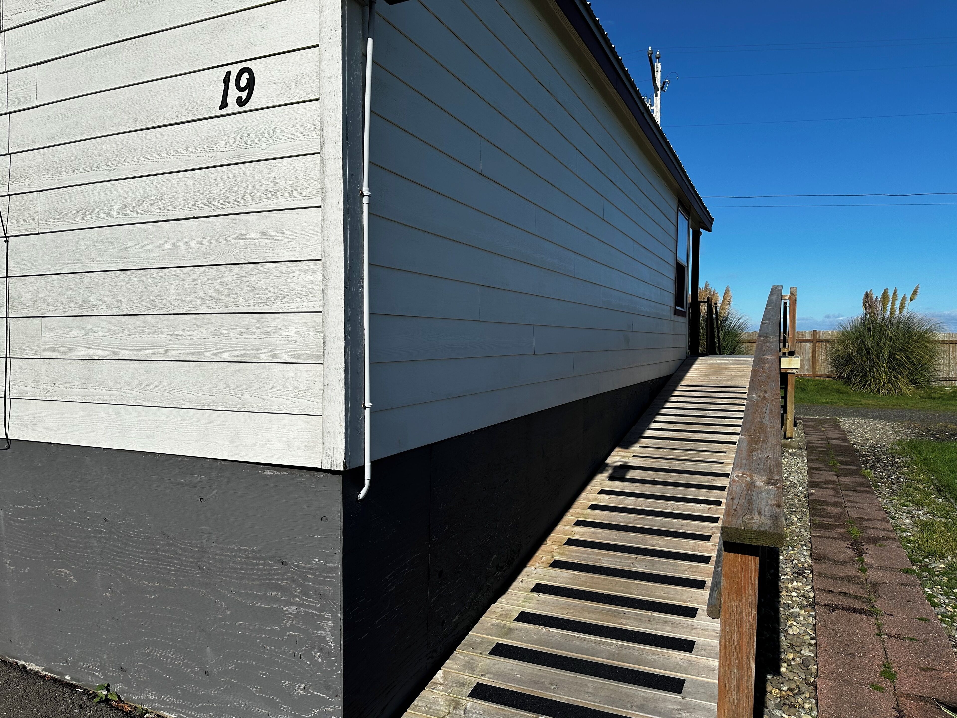 Un bâtiment portant le numéro 19 sur sa façade, une rampe en bois menant à l’entrée, et un ciel bleu clair en arrière-plan.
