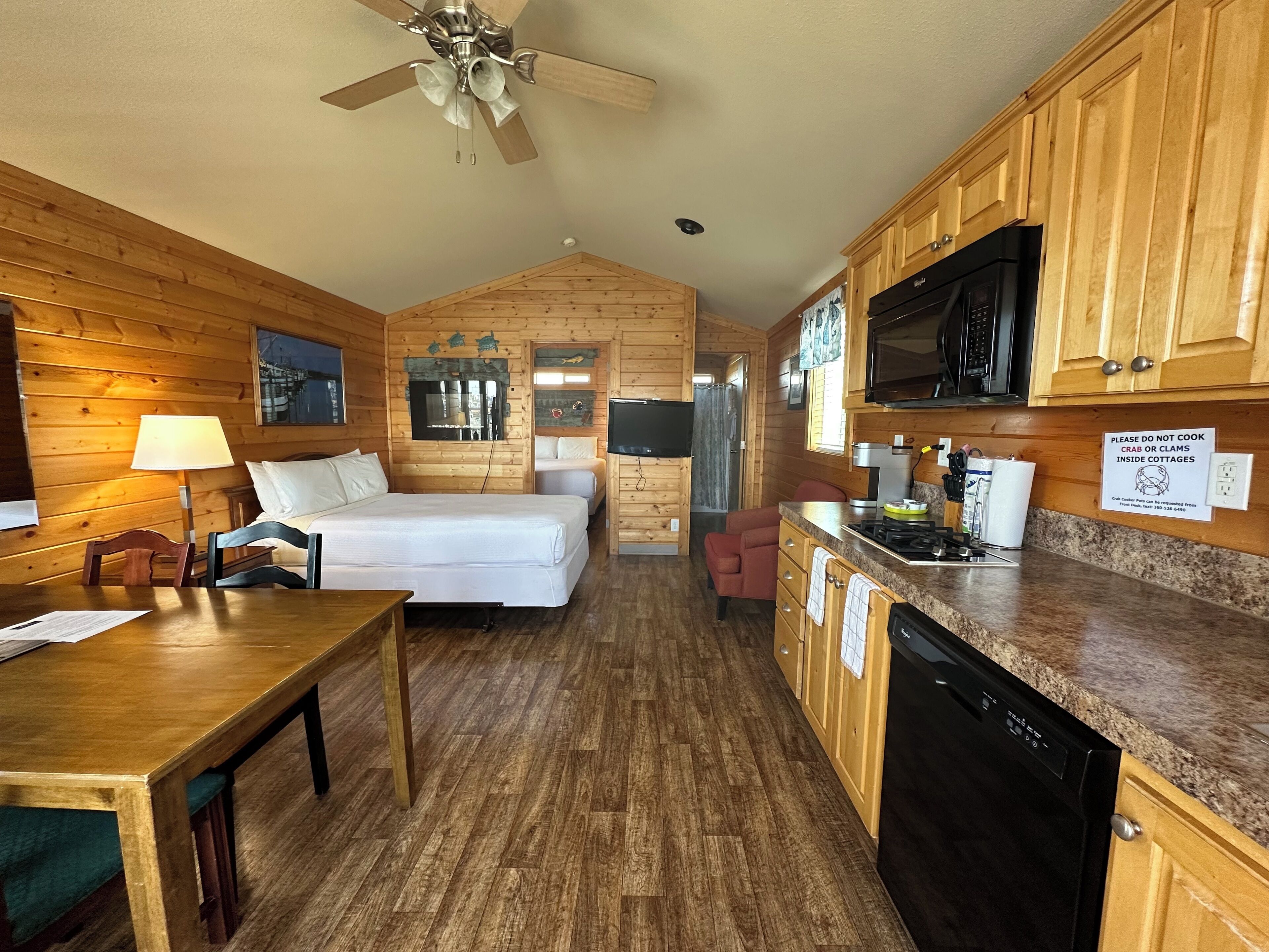 A compact cabin-style room with a bed, a desk, a dining table, a kitchenette, and a ceiling fan.
