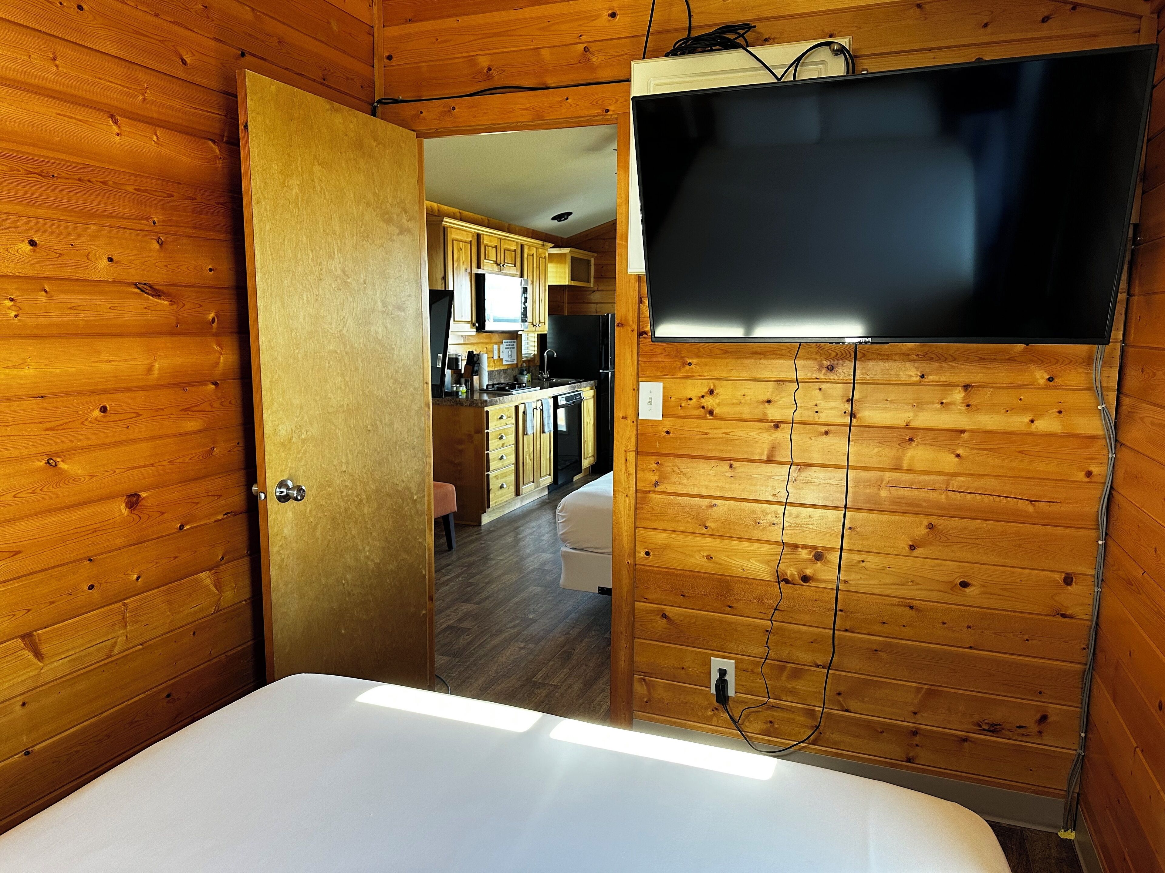 A wooden-paneled room with a bed, a TV mounted on the wall, and a kitchen visible through an open door.