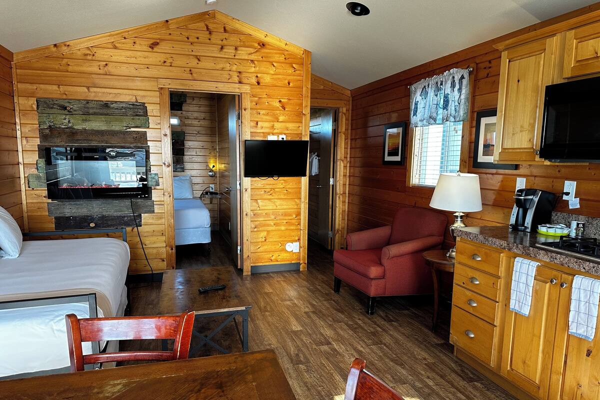 A cozy cabin-style room with a bed, a dining table, a red armchair, a TV, and a kitchenette.