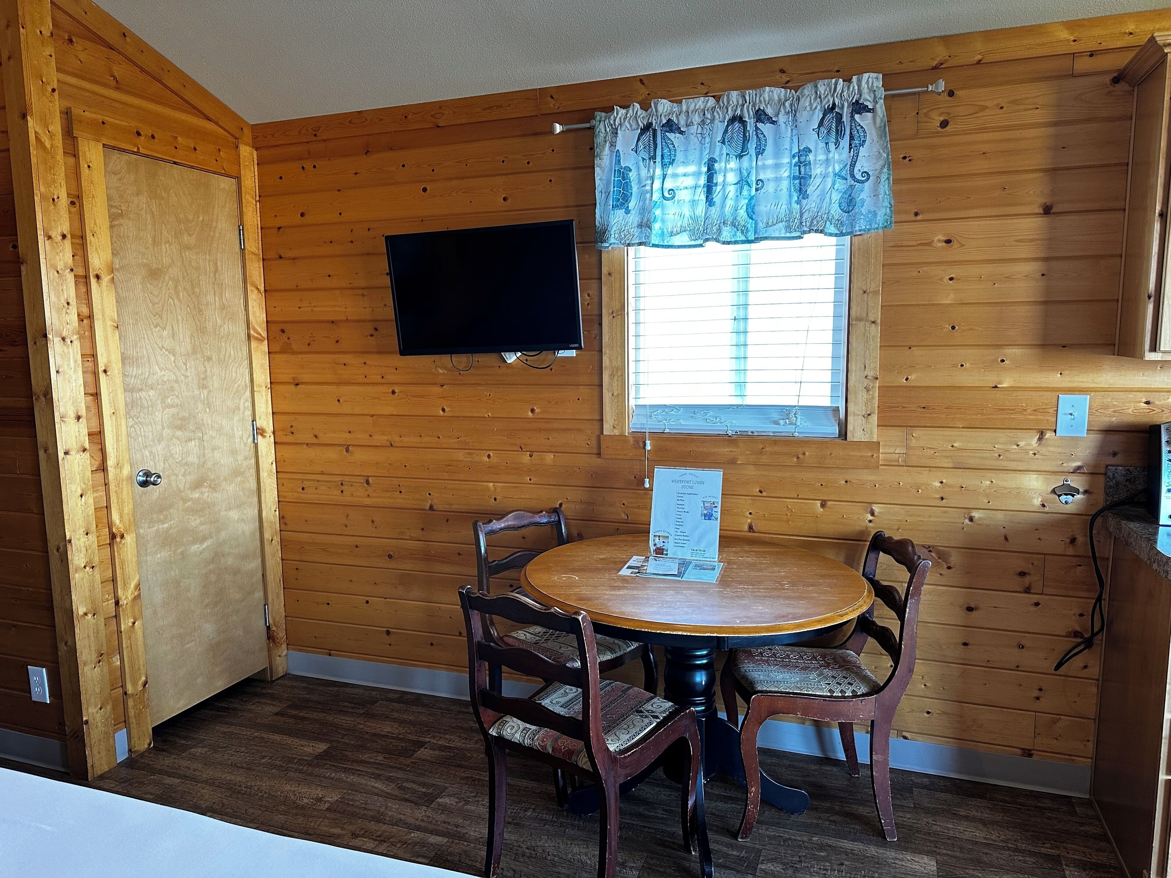 A wooden cabin interior with a round table, four chairs, a television mounted on the wall, and a window with a curtain.