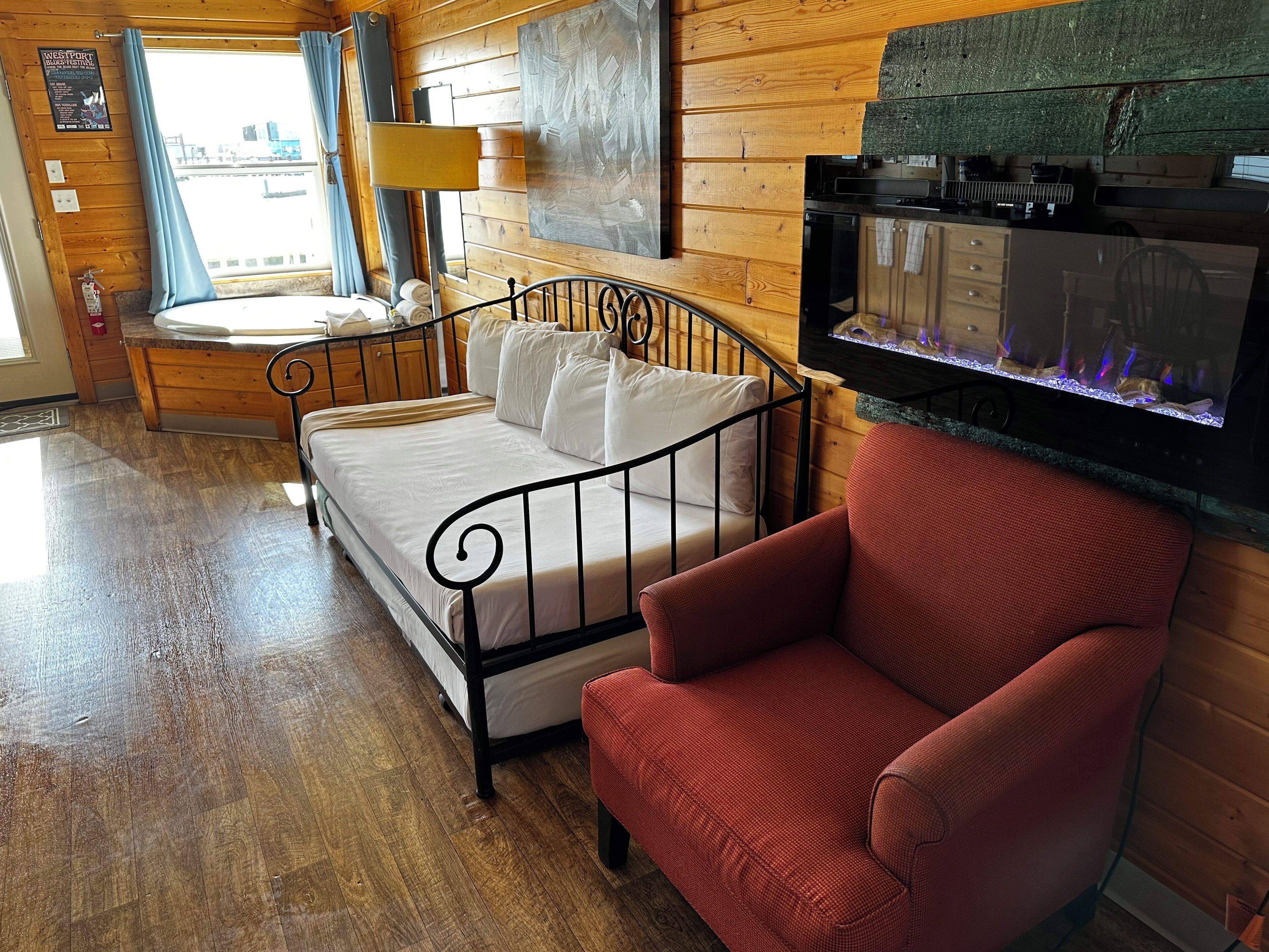 A wooden cabin room with a bed, a red chair, a fireplace, and a window with curtains.