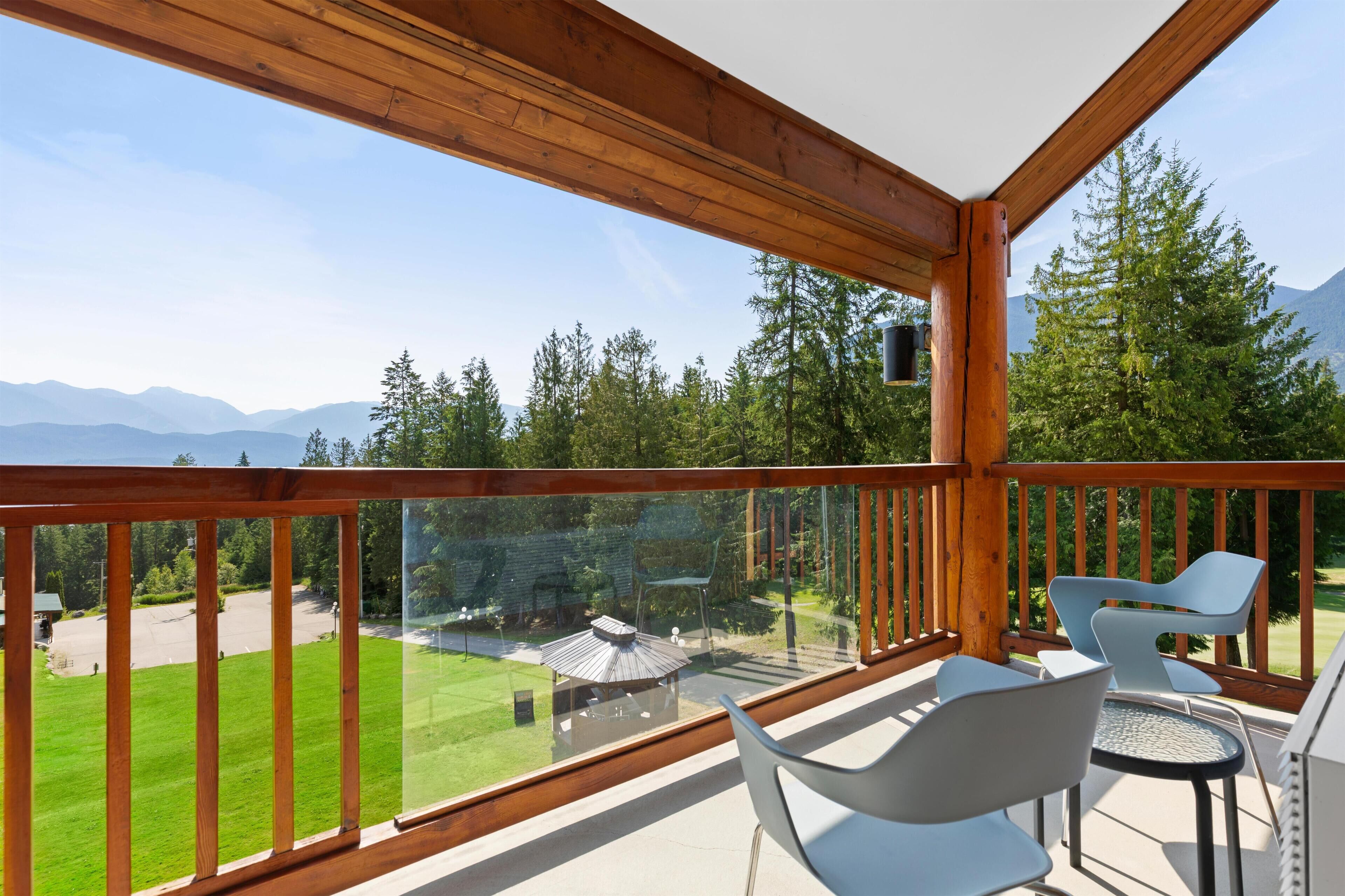 A balcony with wooden railings, a table, and two chairs, overlooking a green landscape and mountains.