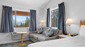 Une chambre d’hôtel avec un lit, un canapé, une petite table, deux lampes et une vue sur les montagnes, visible à travers de grandes fenêtres.