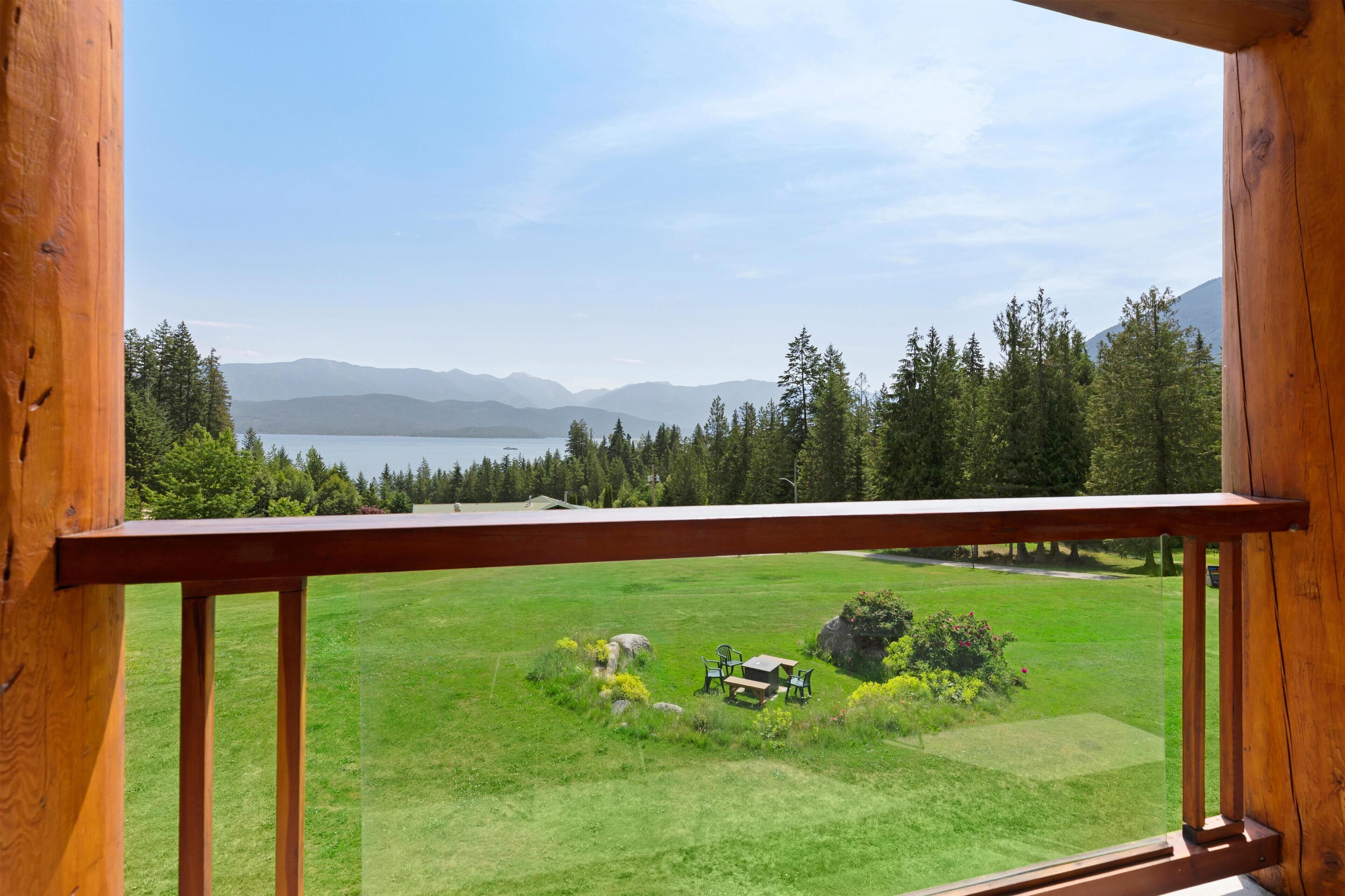 A wooden balcony with a view of a green field, a lake, and mountains in the distance.