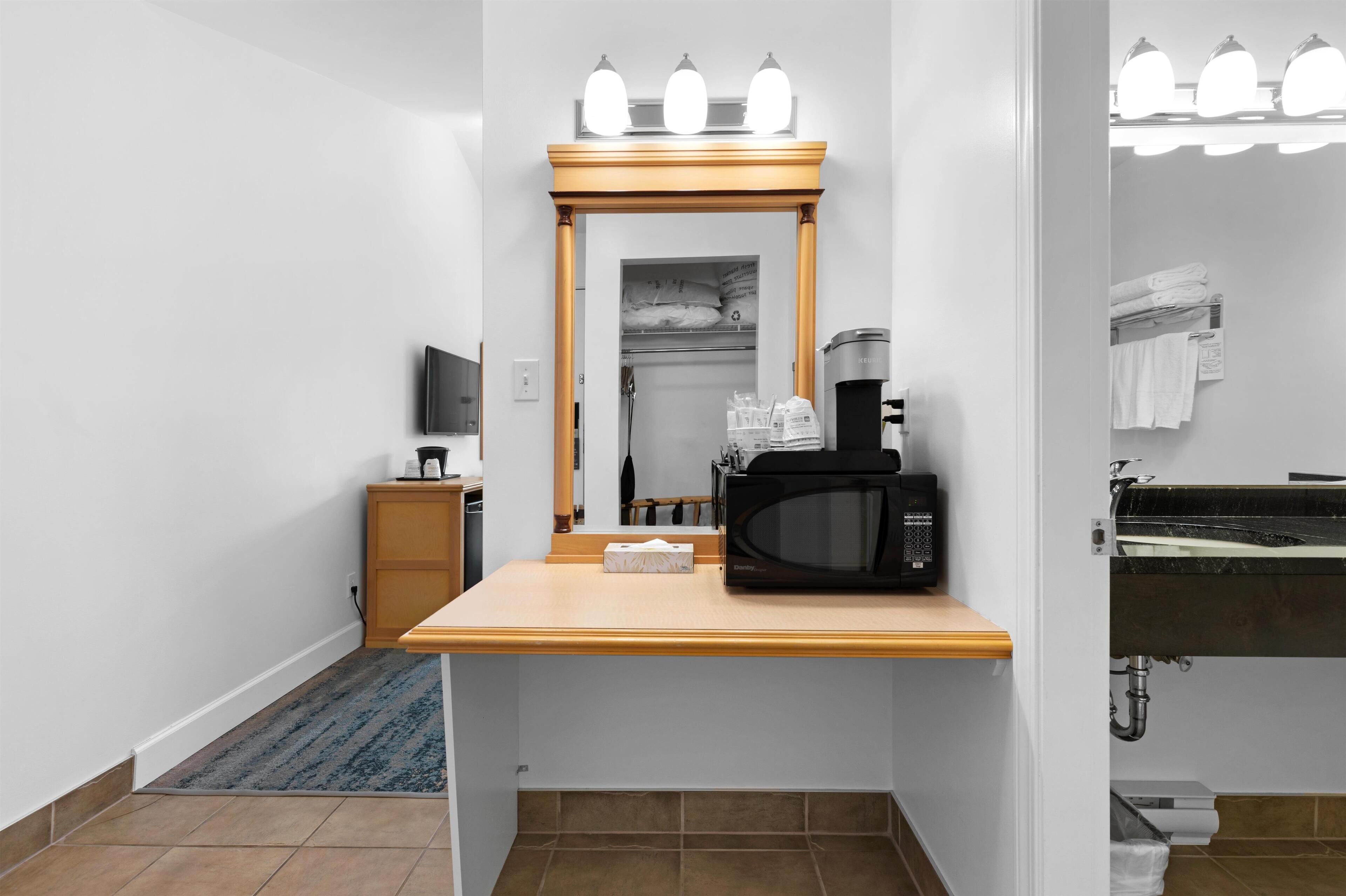 A hotel room with a wooden vanity, a microwave, a coffee maker, a mirror with lights, a small TV, and a bathroom with a sink and towel rack.
