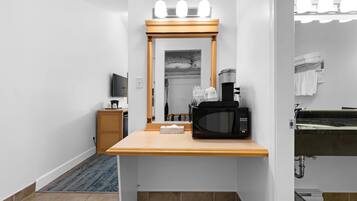 A hotel room with a wooden vanity, a microwave, a coffee maker, a mirror with lights, a small TV, and a bathroom with a sink and towel rack.
