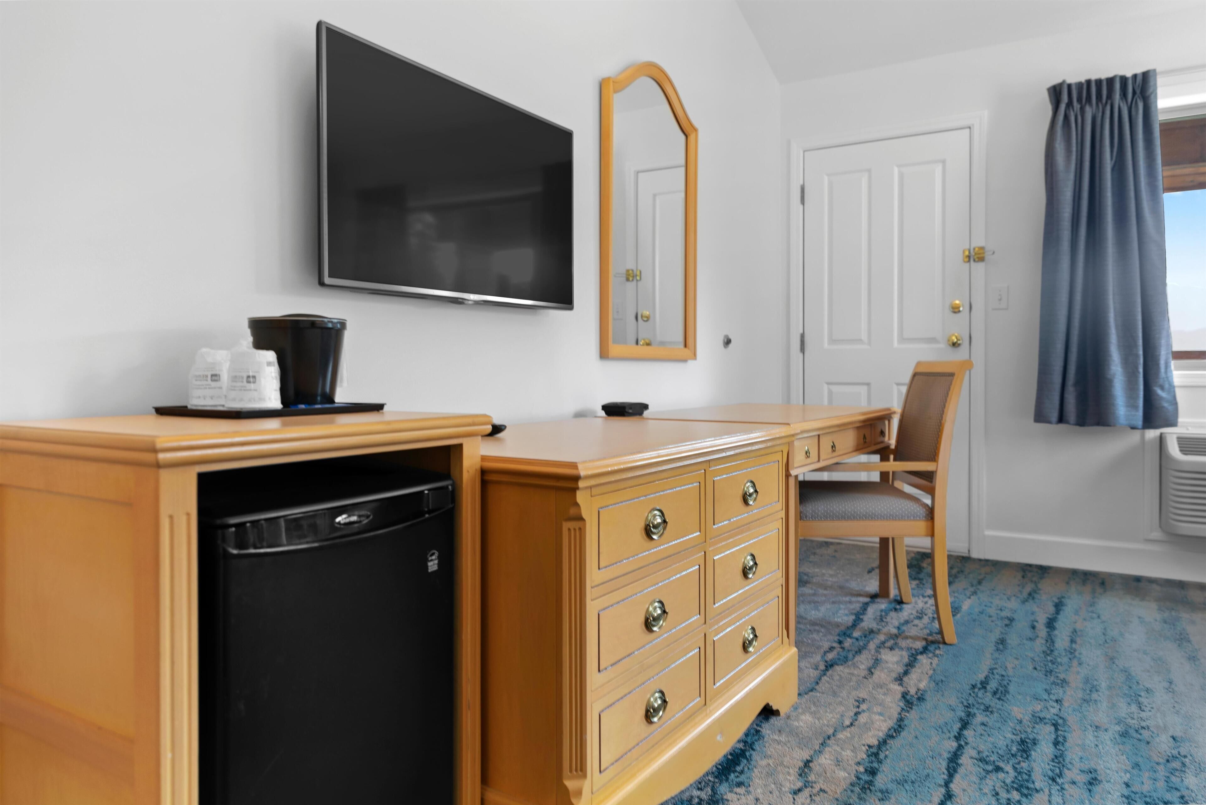 A hotel room with a wooden desk, a television, a mirror, a chair, a wardrobe, and a blue carpet.