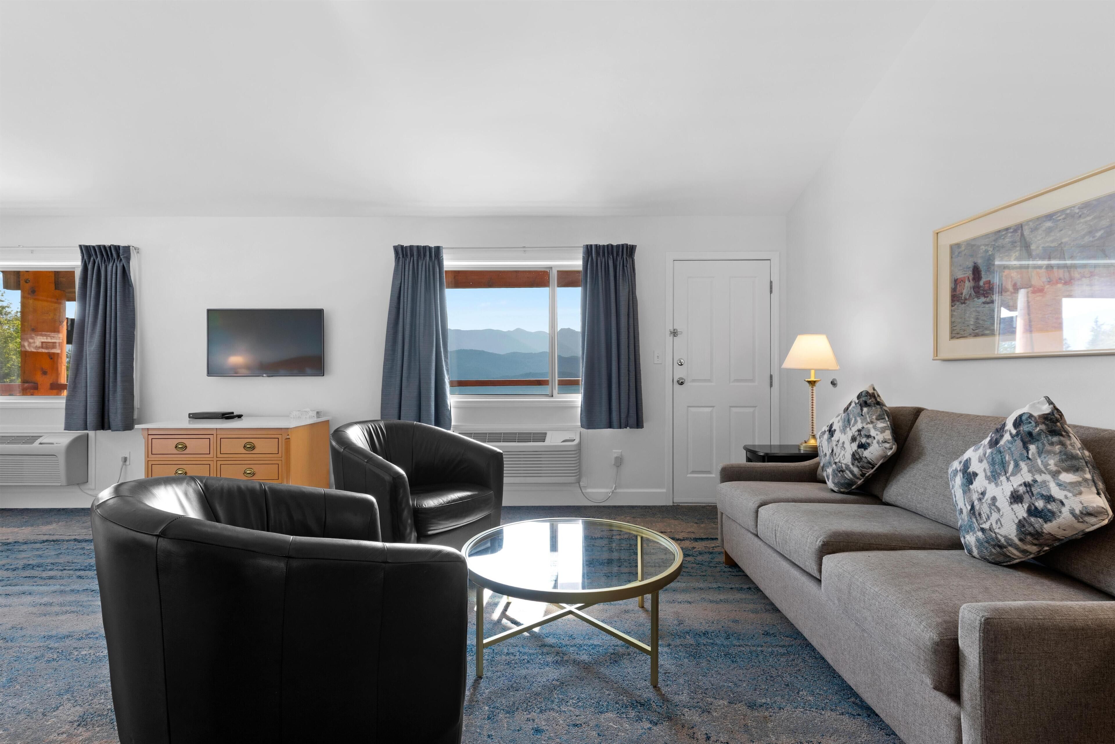 Habitación de hotel con sofá, dos sillones, una mesa de centro de vidrio, televisor, lámpara y un cuadro en la pared.