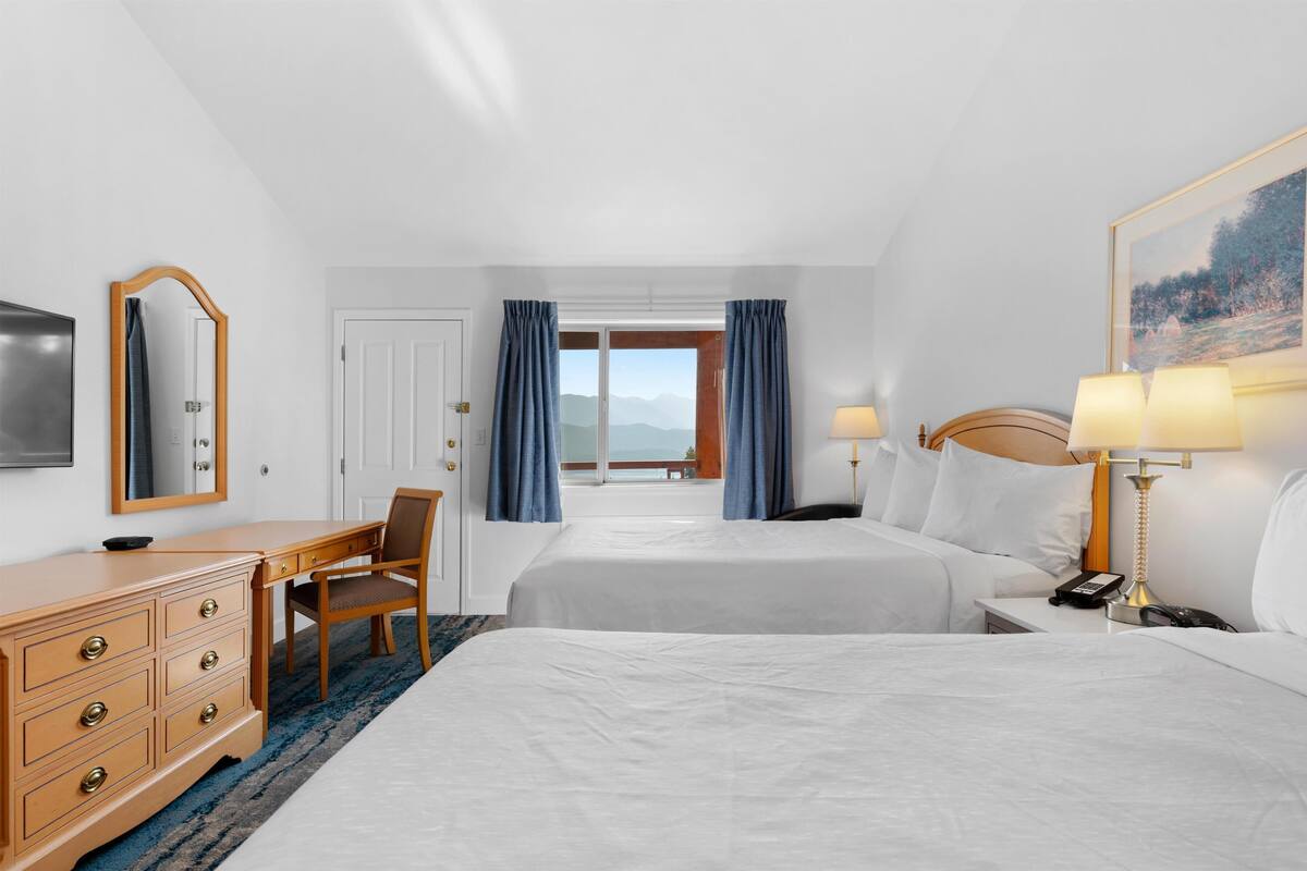 Une chambre d’hôtel avec deux lits, un bureau, une chaise, une commode, un téléviseur, une fenêtre avec des rideaux et un tableau au mur.