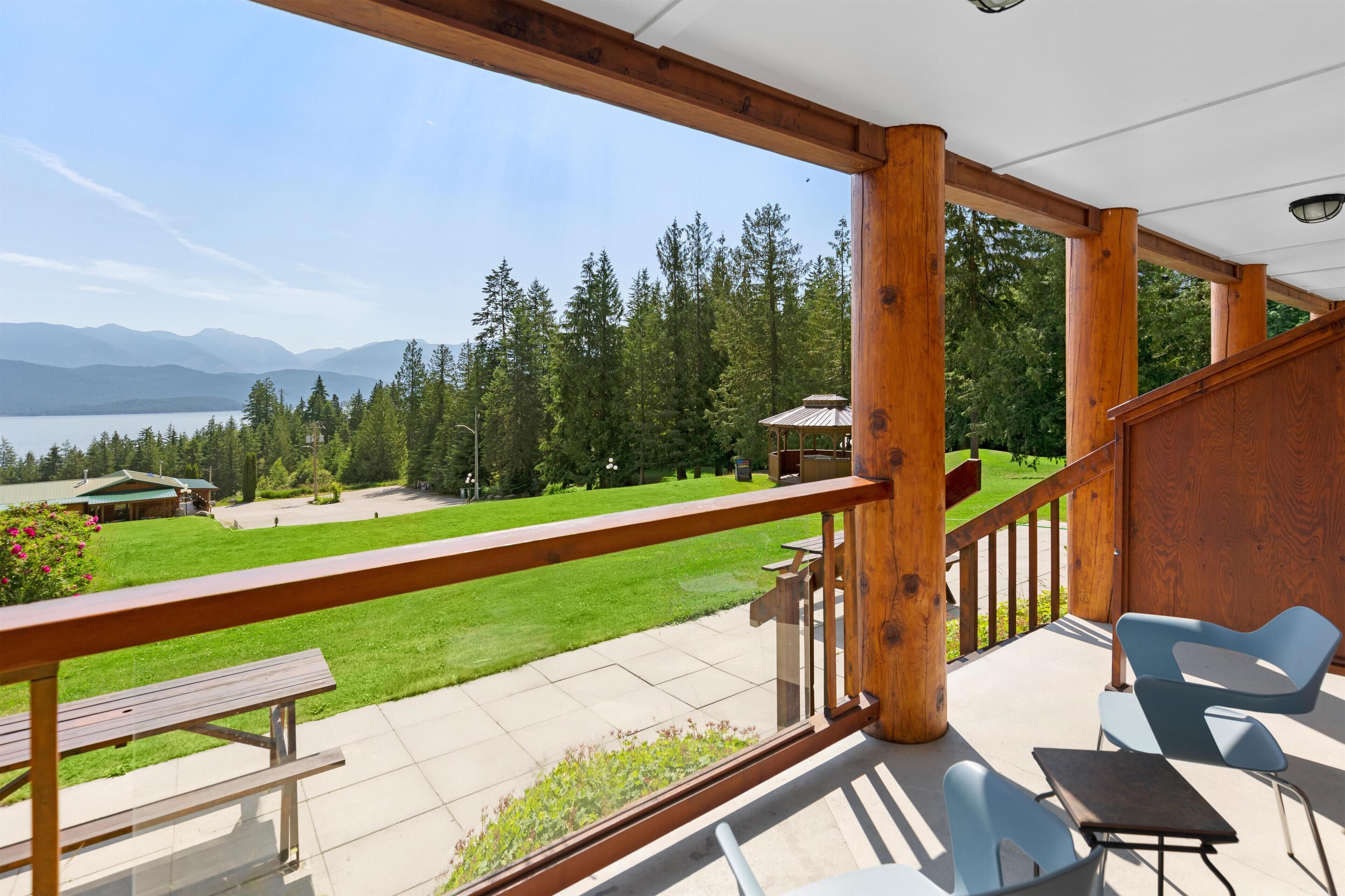 A balcony with wooden railings, a table, and chairs, overlooking a grassy area with trees and a distant mountain range.