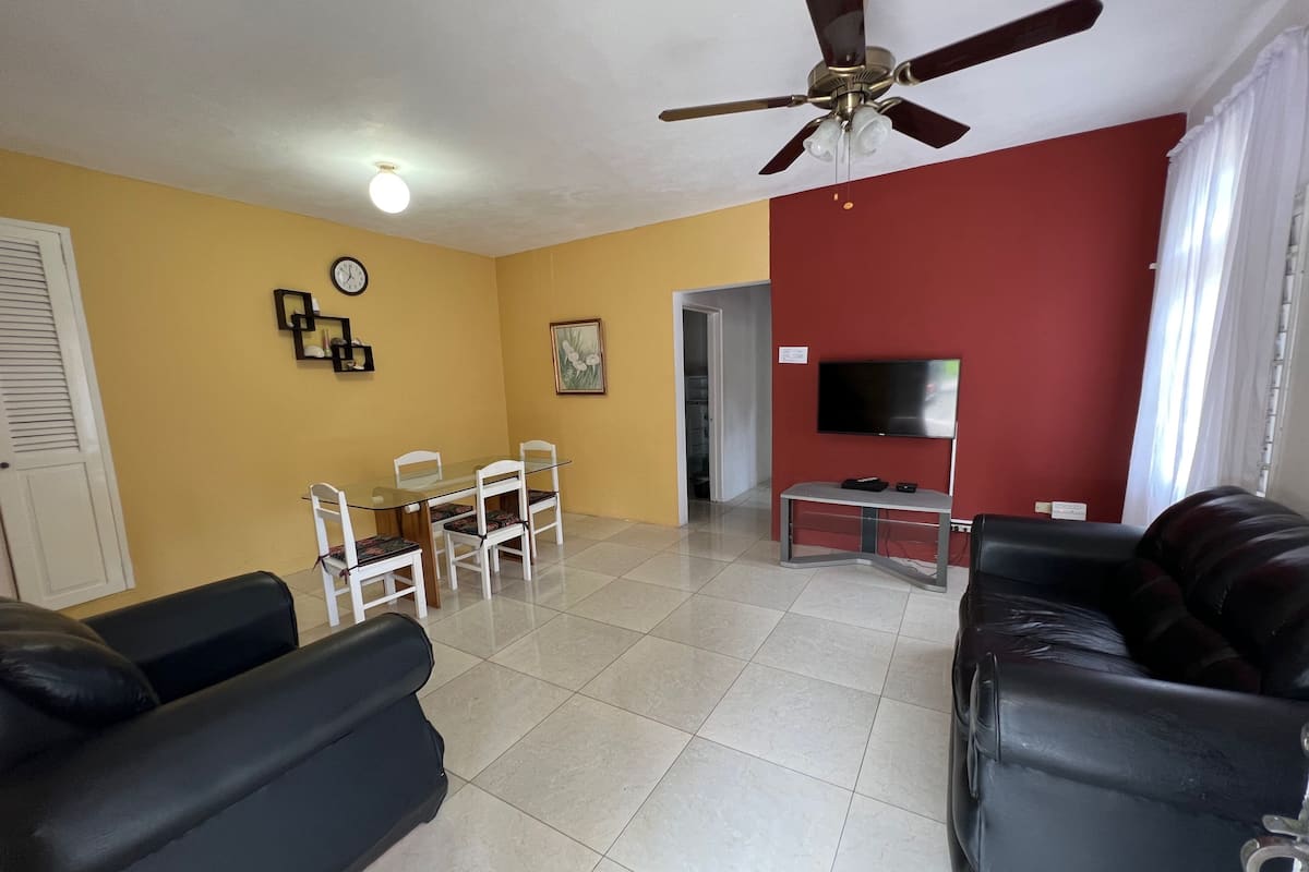 A living room with a ceiling fan, a dining table with chairs, a TV on a stand, and two black sofas.
