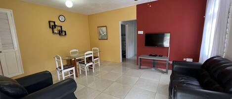 A living room with a ceiling fan, a dining table with chairs, a TV on a stand, and two black sofas.