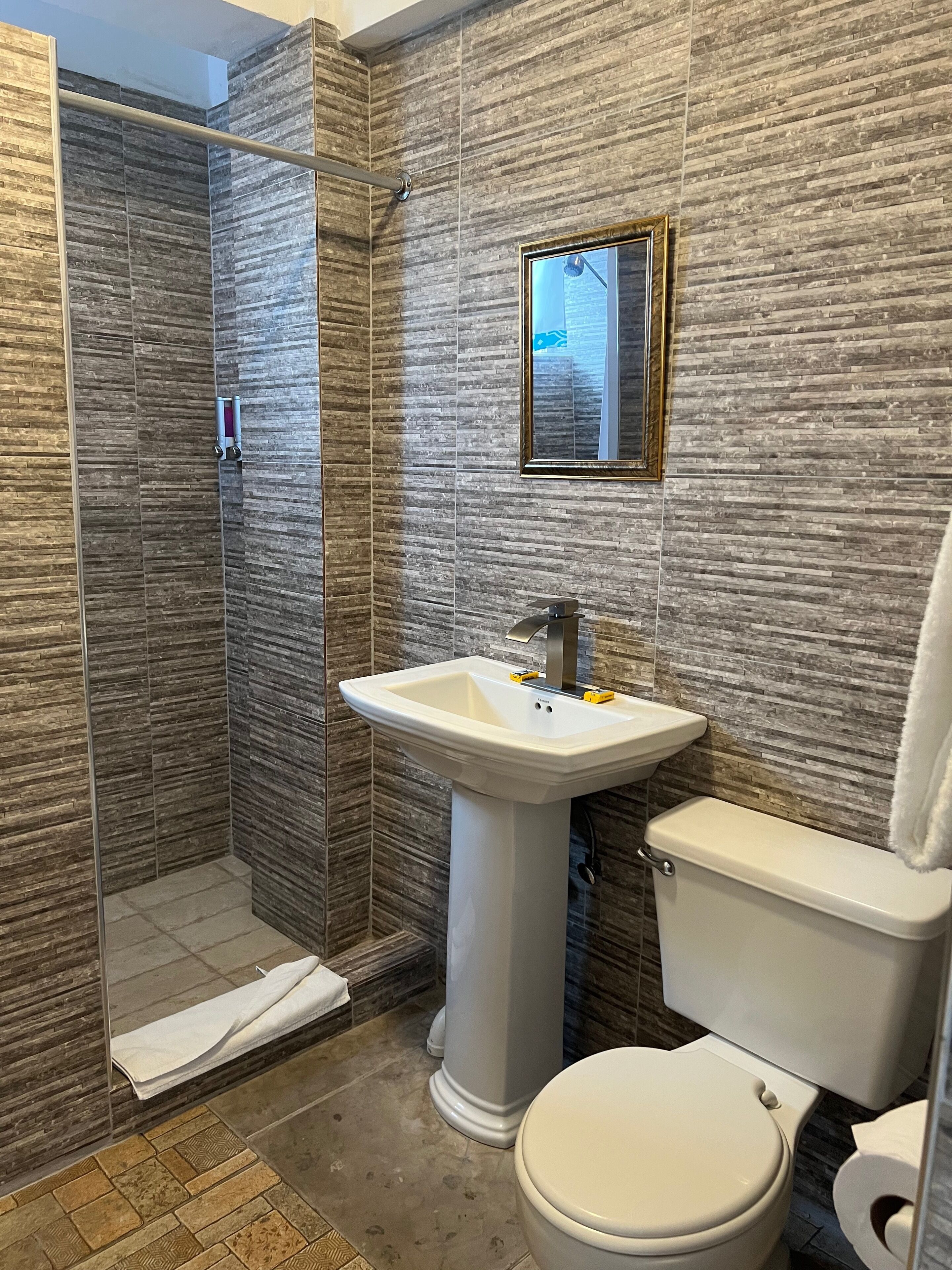 A bathroom with a shower, sink, and toilet, featuring textured wall tiles and a framed mirror.