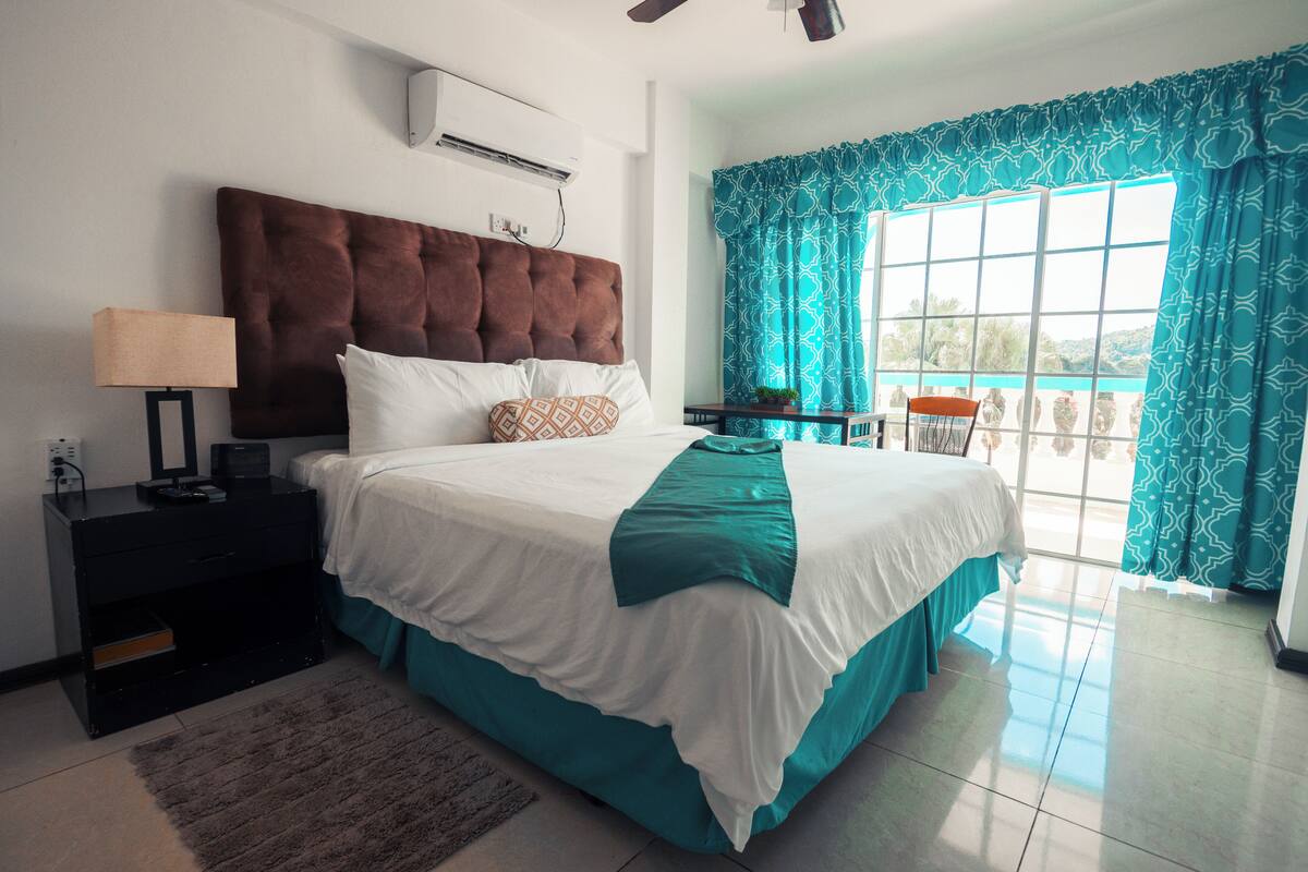 A bedroom with a large bed, a nightstand, a lamp, and a view of the outdoors through a window with curtains.