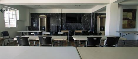 Un aula con mesas y sillas, un televisor en la pared y una ventana.