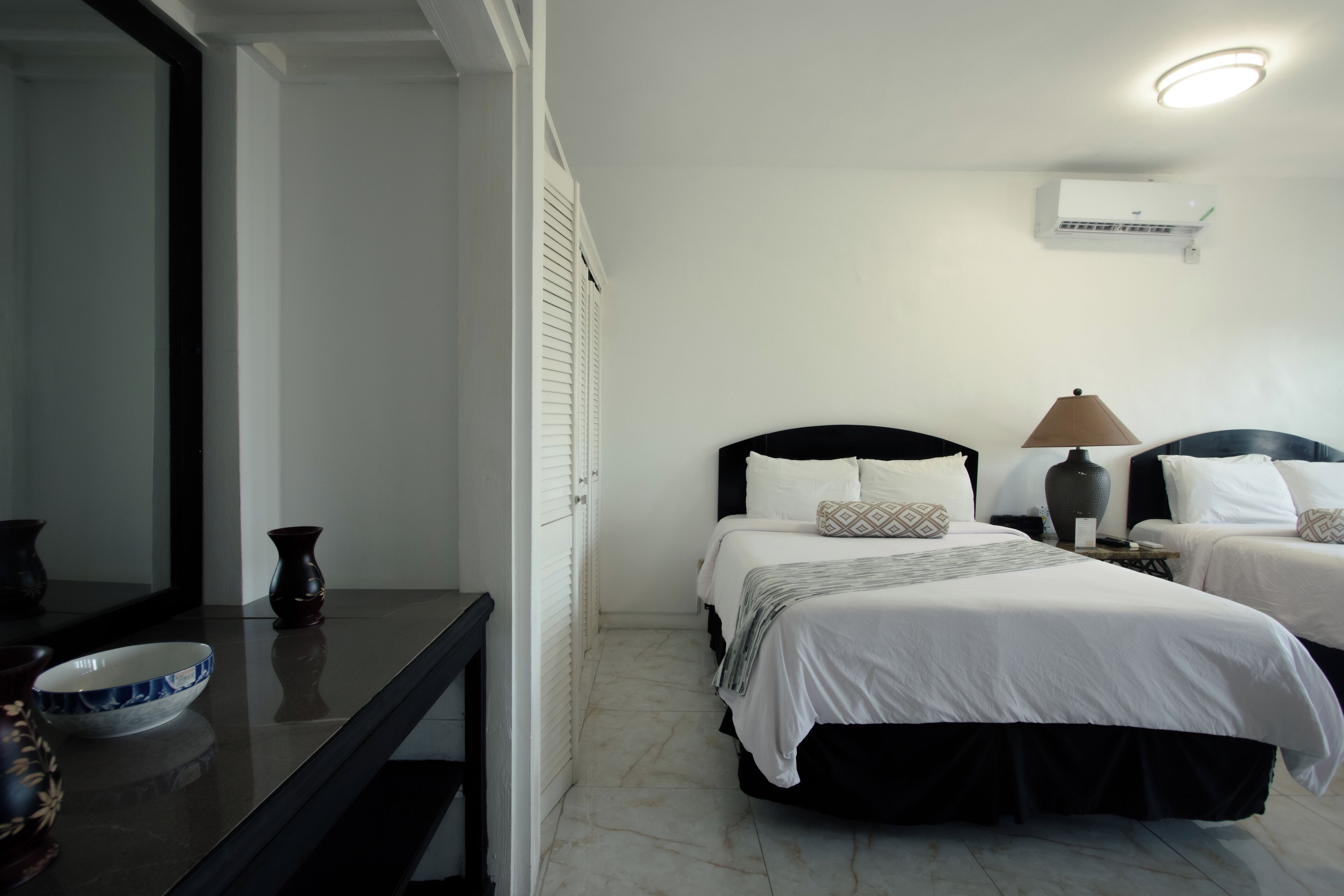 A hotel room with two beds, a bedside table with a lamp, a mirror, and a bowl on the table.