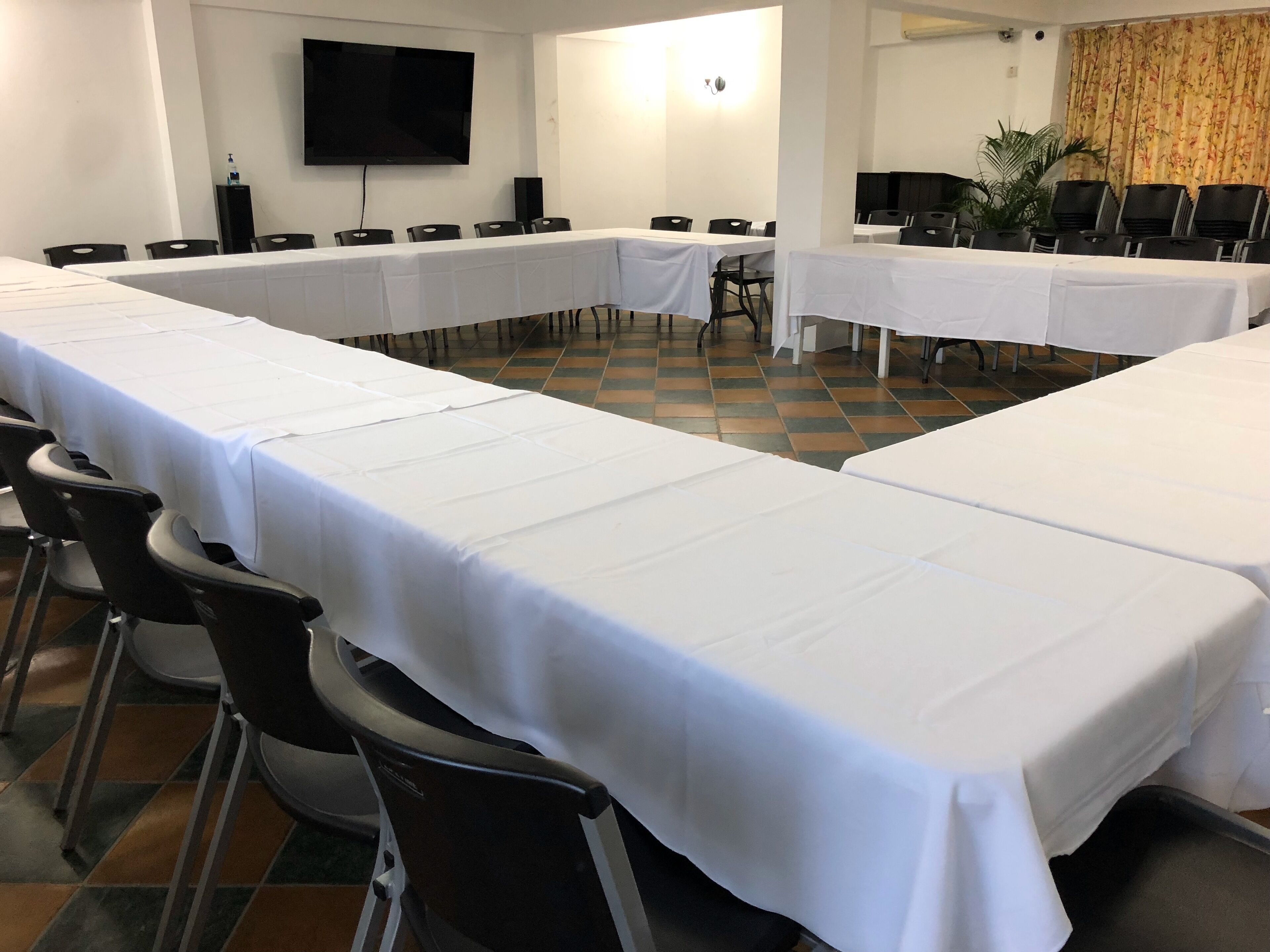 Un salón de conferencias con largas mesas cubiertas con manteles blancos, sillas negras, un televisor de pantalla plana en la pared y una planta en maceta.