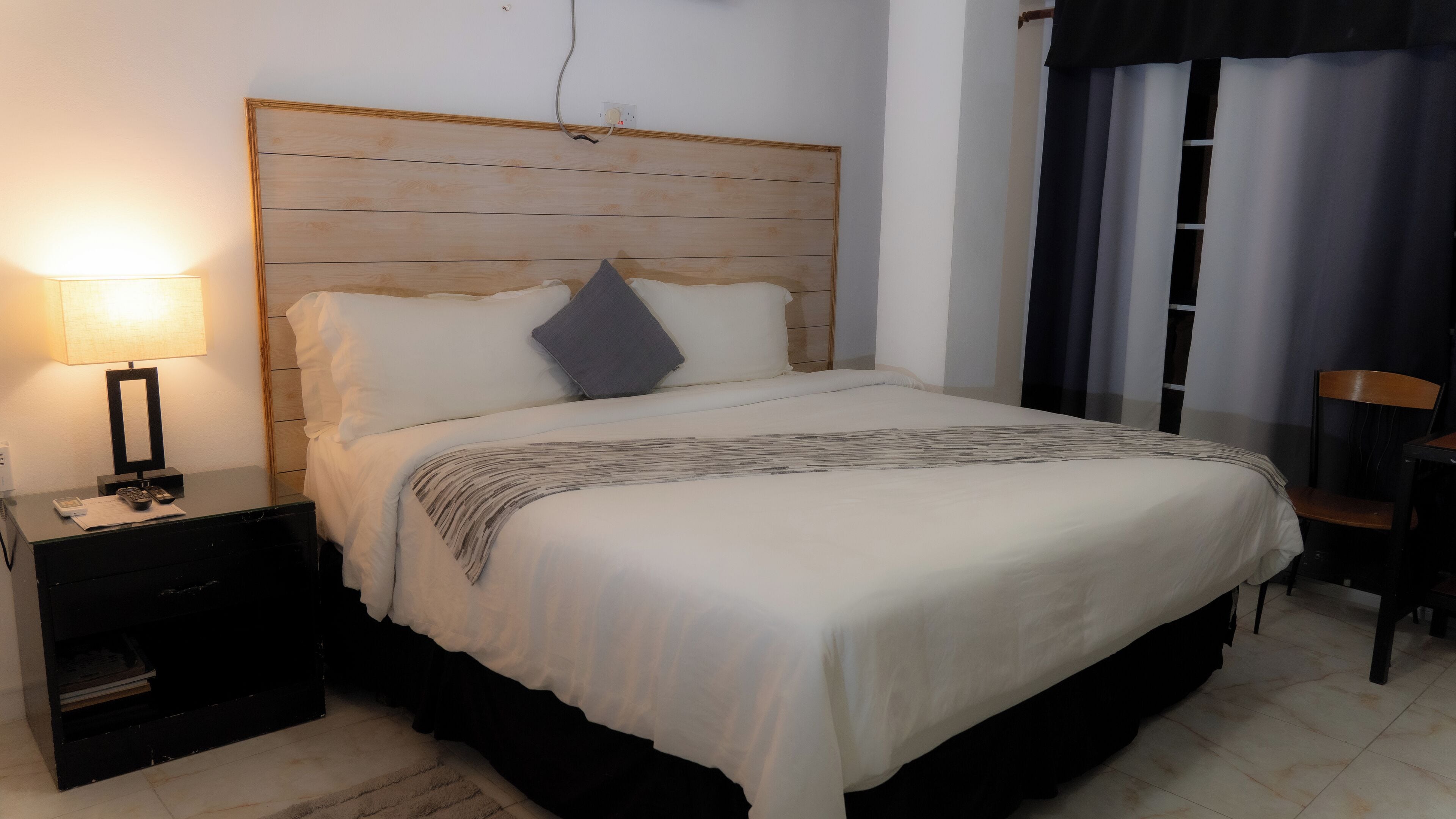 Ein ordentlich bezogenes Bett mit Holz-Kopfteil, einer Nachttischlampe und einem Sessel in einem Zimmer mit Fliesenboden.