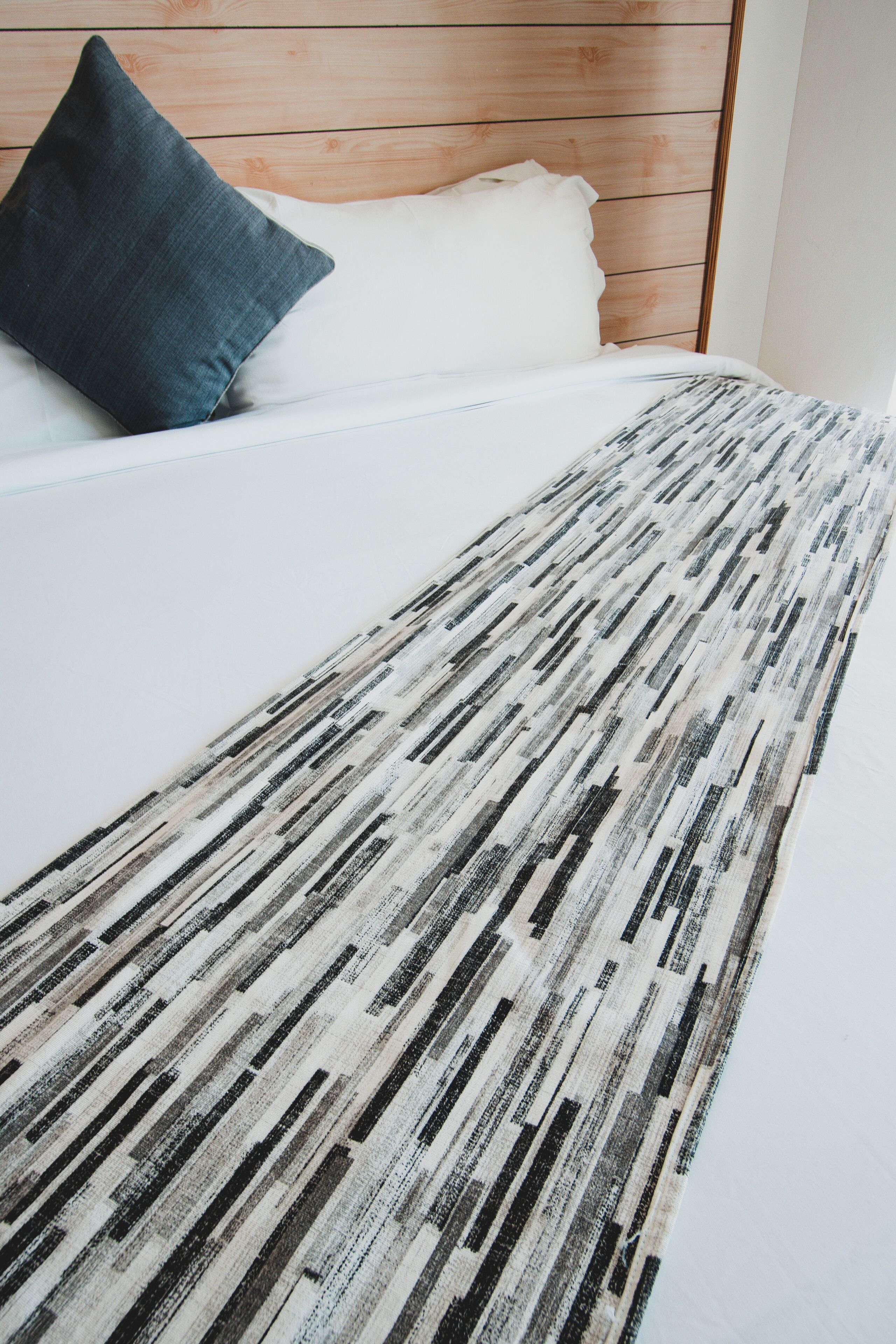 Ein Bett mit weißer Matratze und einem dunkelblauen Kissen, ein Teppich im weißen und schwarzen Streifenmuster sowie ein Holzkopfbrett.