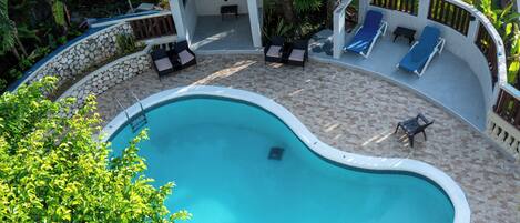 A swimming pool with a curved edge, surrounded by a stone wall and a tiled deck. There are lounge chairs and a small table near the pool.