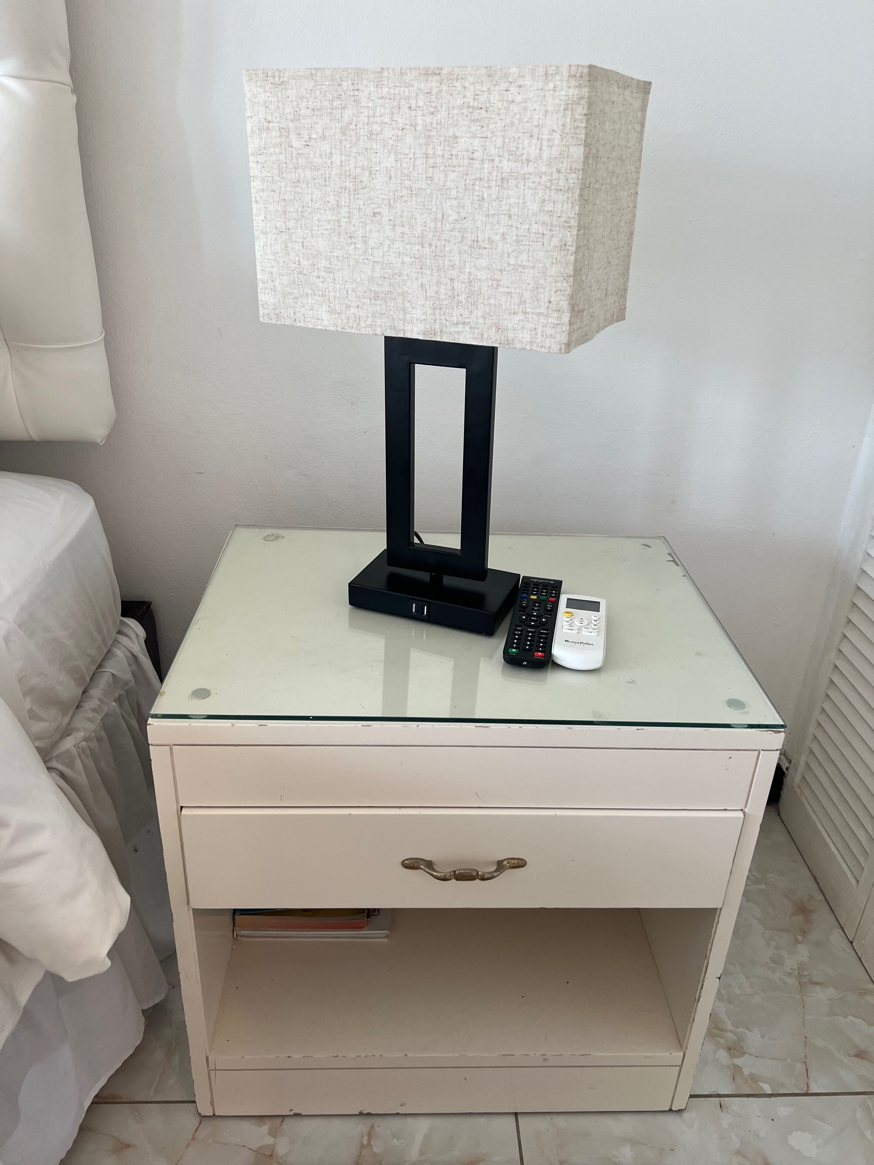 A white bedside table with a lamp, a remote control, and a mobile phone.