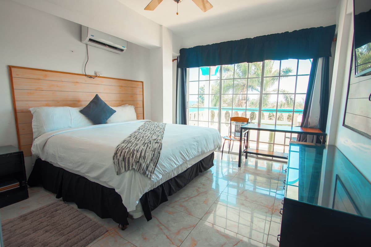 A bedroom with a large bed, a wooden headboard, a small table, and a chair near a window with a view of palm trees.