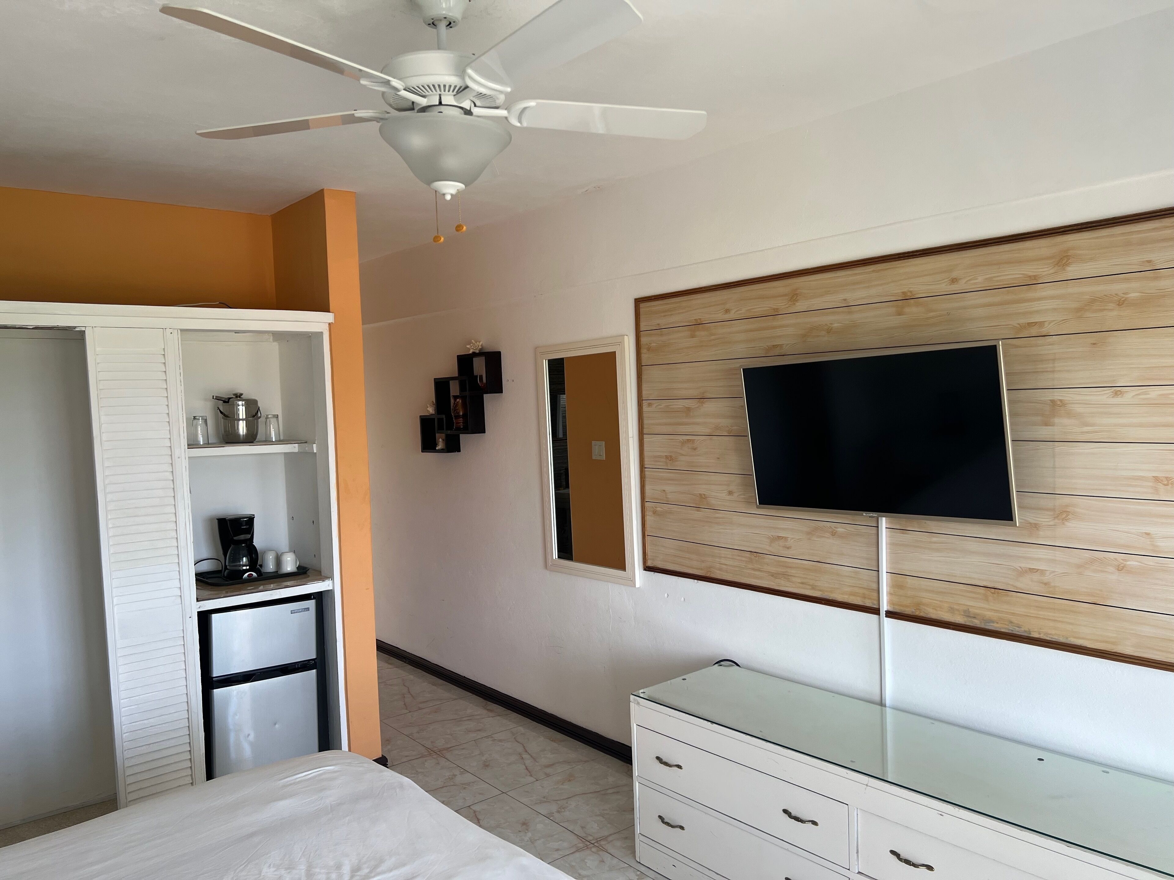 A bedroom with a bed, a wardrobe, a dresser, a ceiling fan, a TV mounted on a wooden panel wall, and a coffee maker on the dresser.