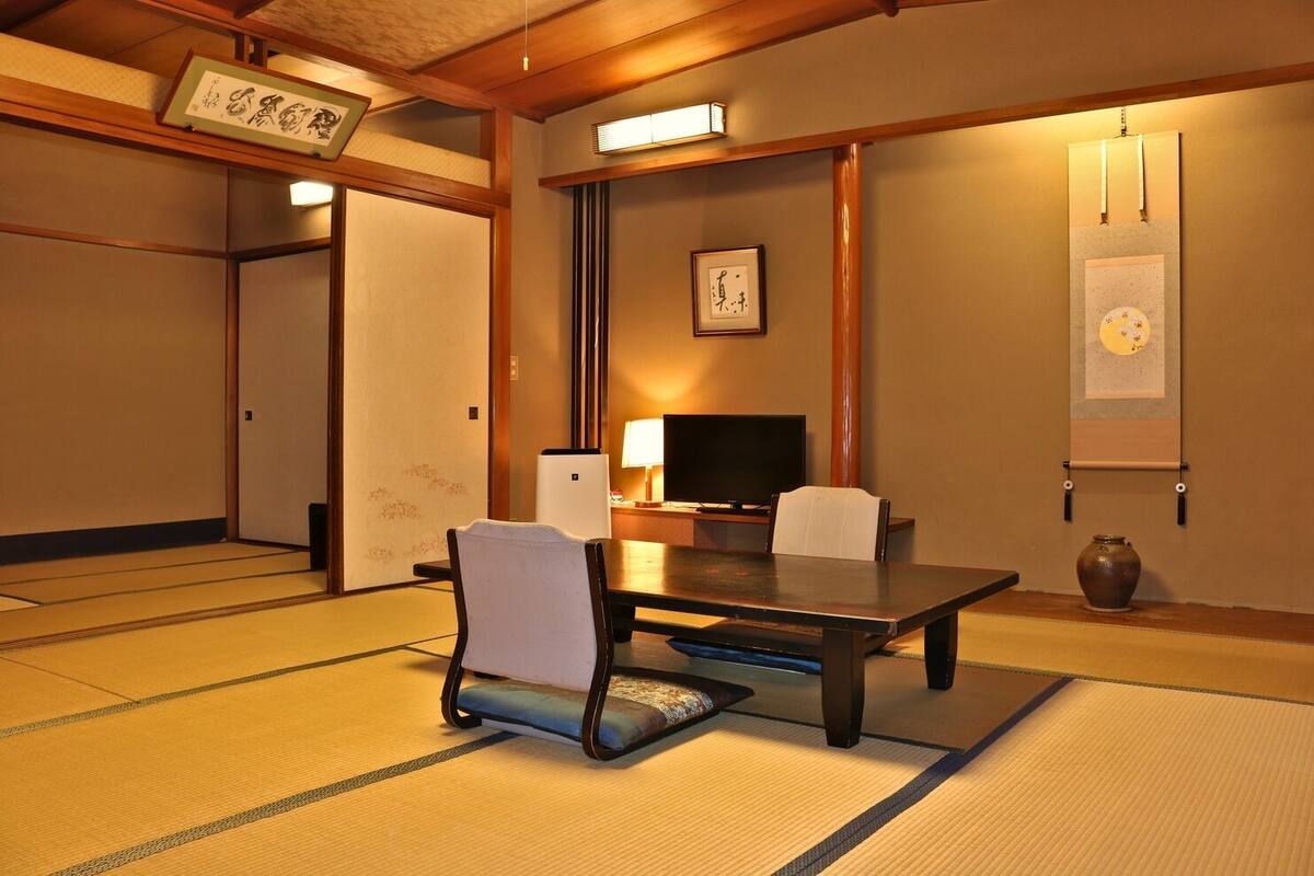 A traditional Japanese room with tatami flooring, a low table, chairs, a television, and artwork on the walls.