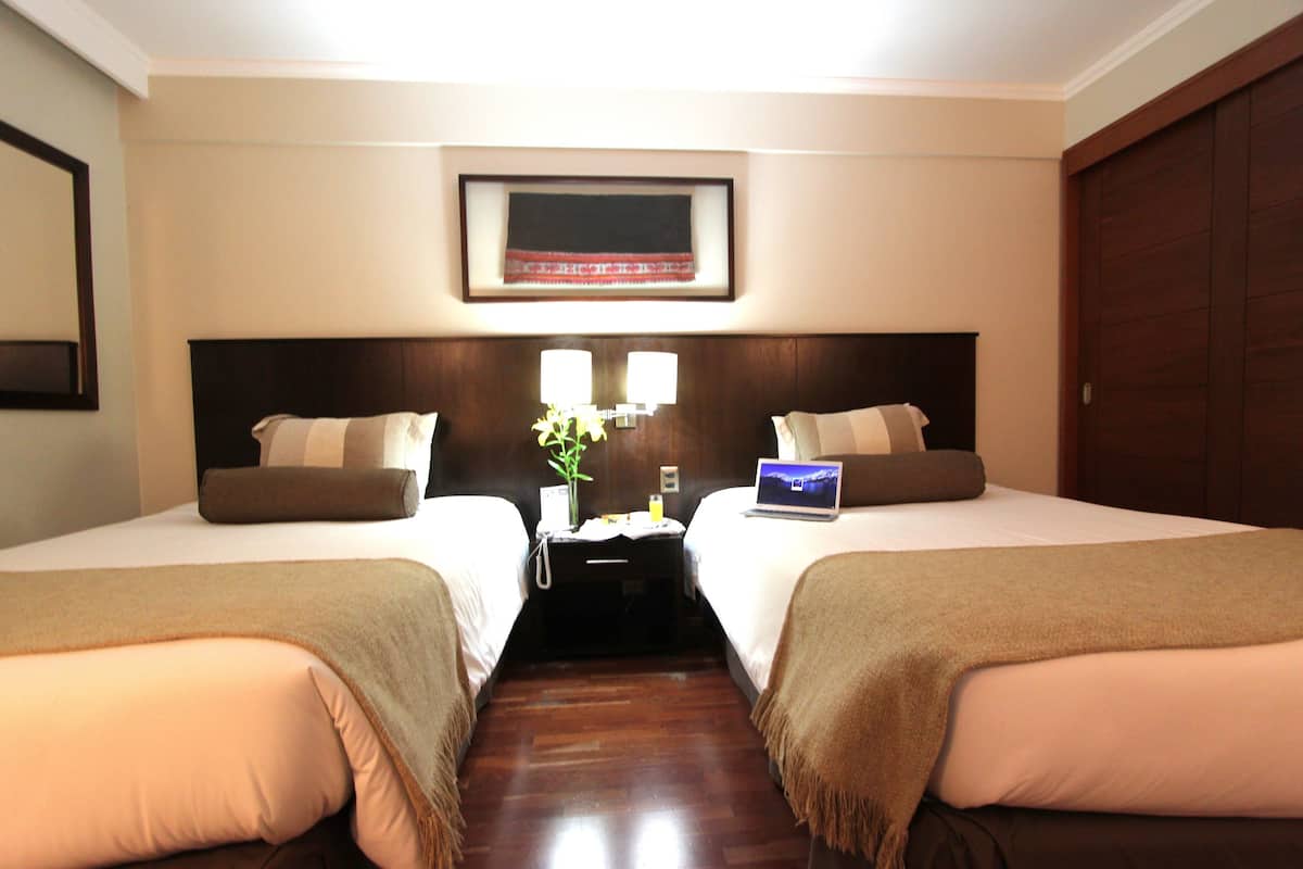A hotel room with two beds, a nightstand with a lamp, a framed picture on the wall, and a wooden floor.