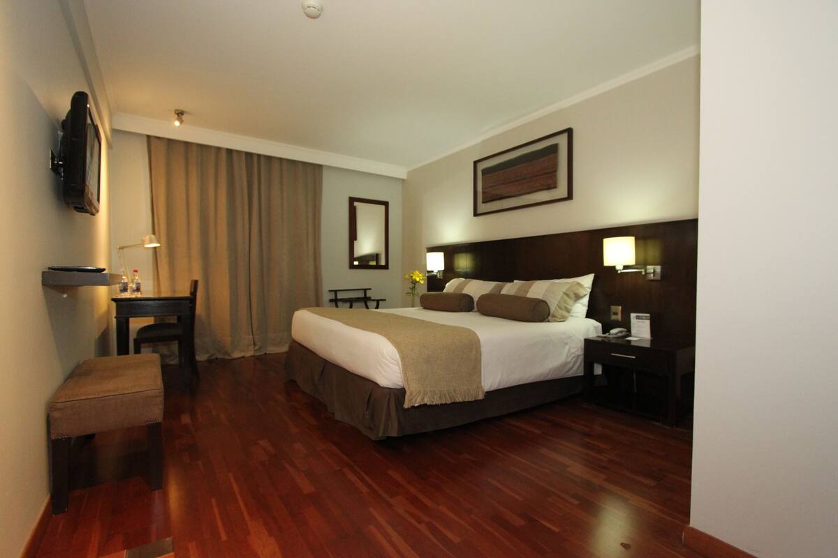 A hotel room with a large bed, a desk with a chair, a television mounted on the wall, and a window with curtains.