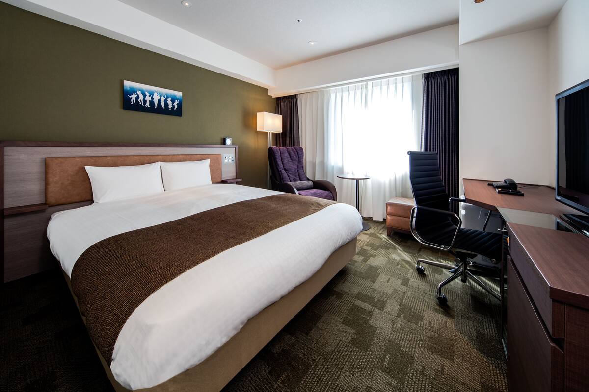 Habitación de hotel con una cama grande, un escritorio con una silla, un televisor y una lámpara.