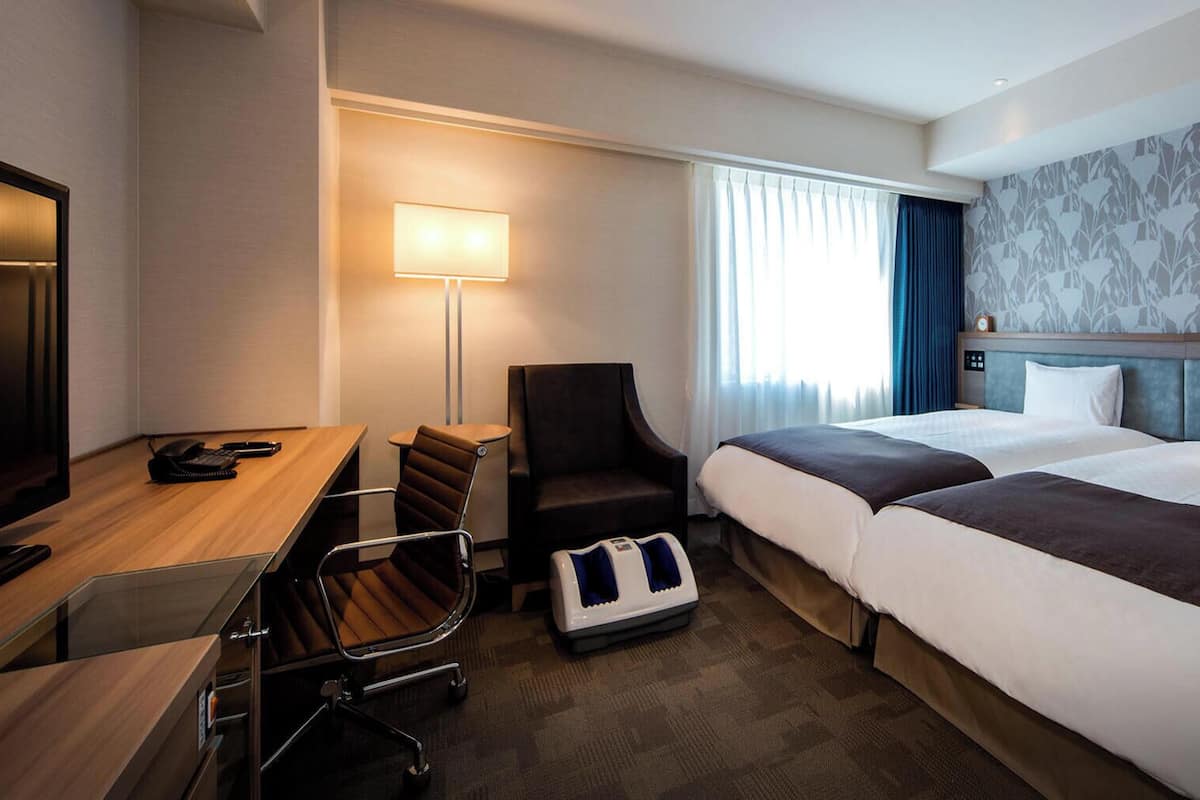 Una habitación de hotel con escritorio, dos camas, una silla, lámpara y ventana con cortinas.