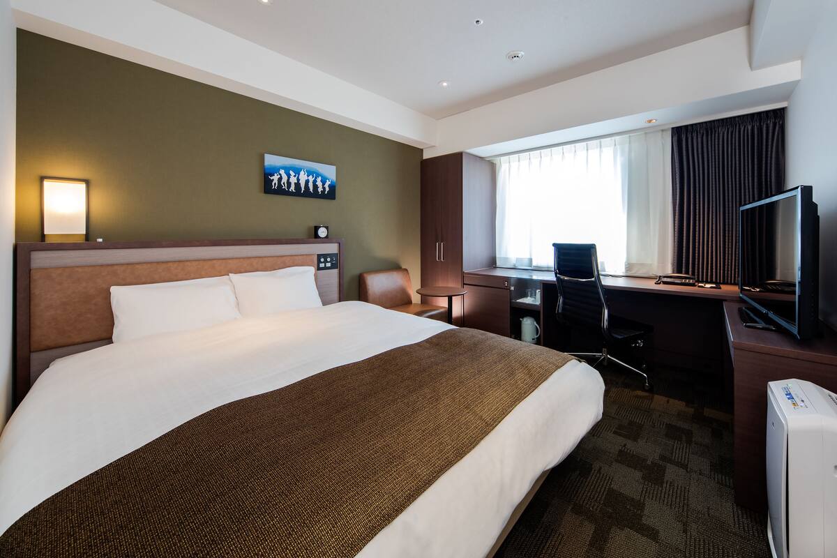 Una habitación de hotel con una cama grande, un escritorio, una silla, un televisor y un ventanal con cortinas.