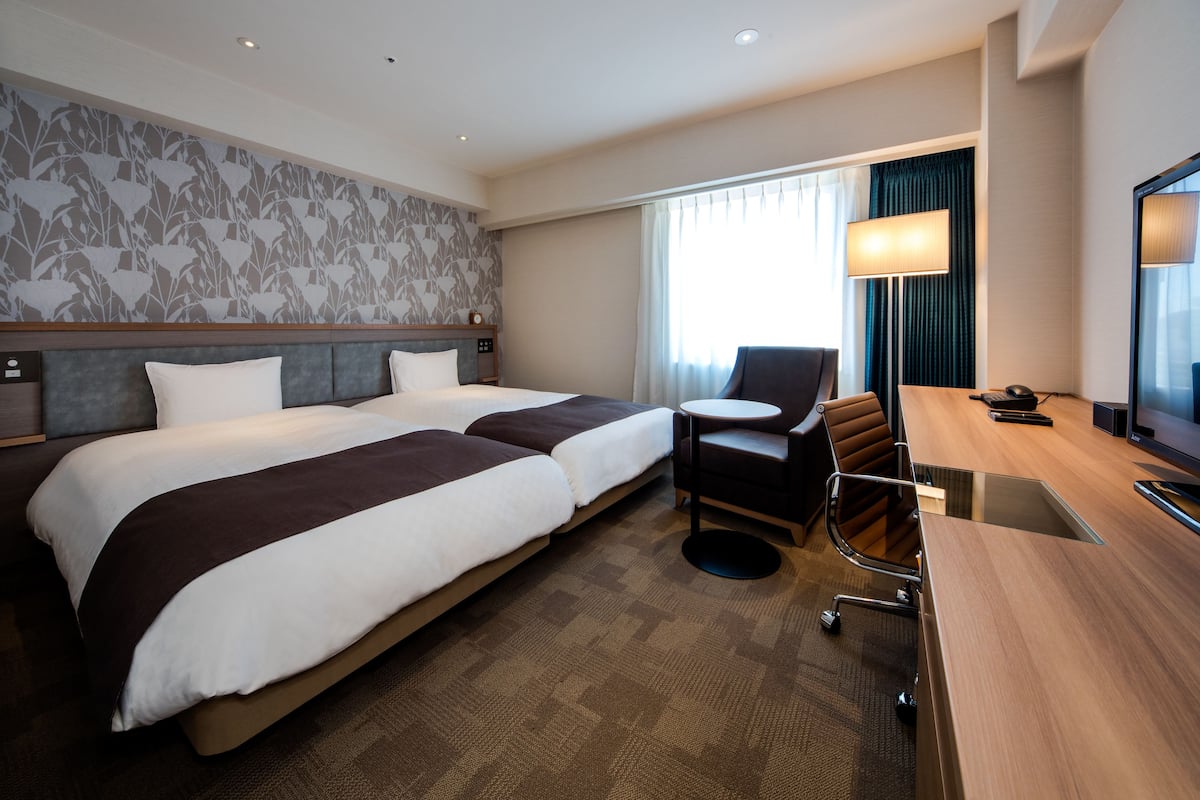 Habitación de hotel con dos camas, un escritorio, una silla, una lámpara y un televisor.