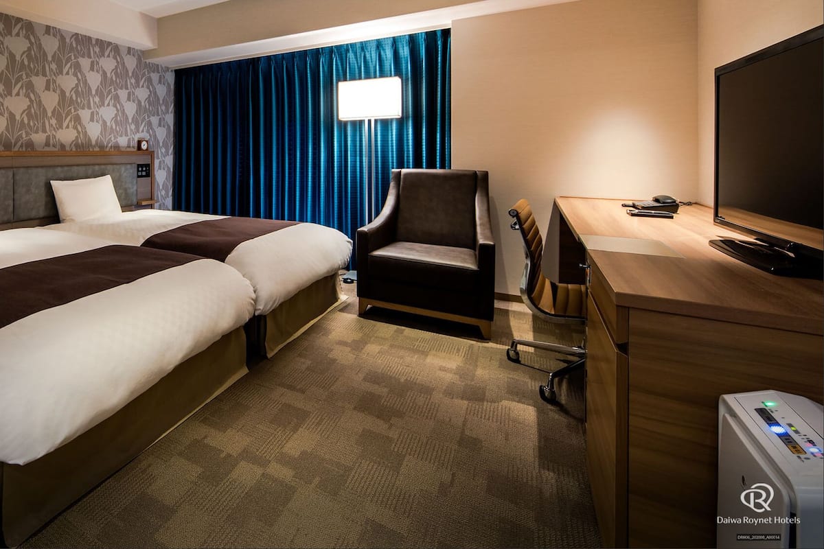 Habitación de hotel con dos camas, un escritorio con computadora, una silla, televisor y ventana con cortinas.