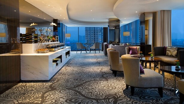 Un moderno vestíbulo de hotel con una recepción de mármol, zona de estar con sofás y sillones, alfombra estampada y grandes ventanales con vistas a la ciudad.