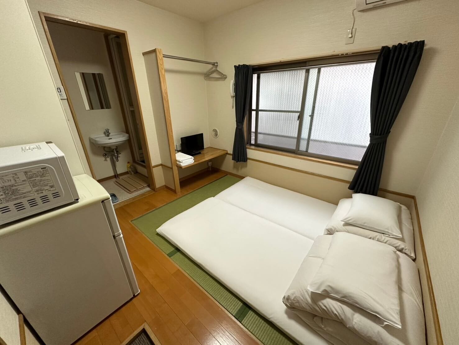 A compact hotel room with a single bed, a small desk with a television, a compact kitchenette, and a bathroom.