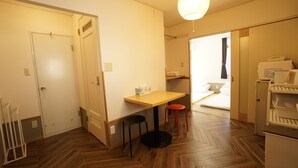 Habitación de hotel compacta con suelo de madera en espiga, una mesita con una silla, kitchenette y cama con ropa blanca.