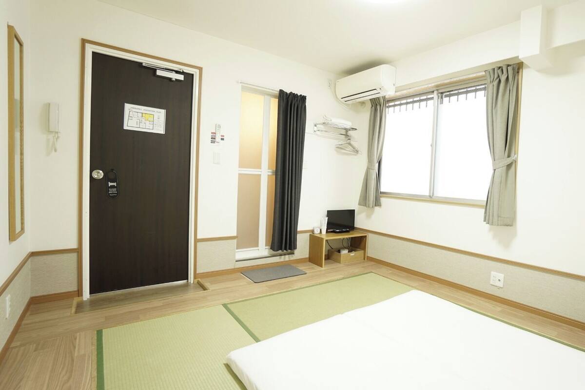 A hotel room with a bed, a TV on a stand, a window with curtains, a door with a sign, and a small table with a remote control.