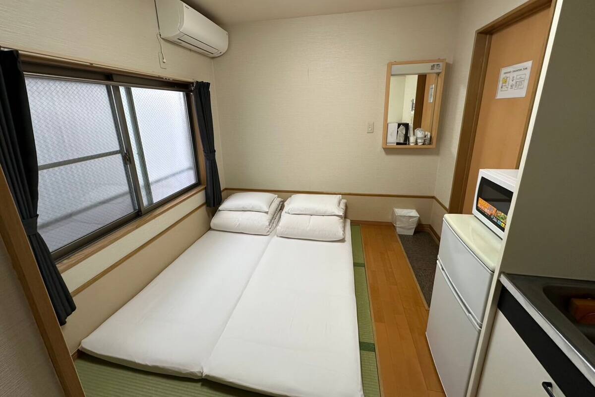 A compact room with a bed, a small kitchenette, a window with curtains, and a mirror.