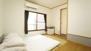A bedroom with a bed, a window with curtains, an air conditioner, a small refrigerator, and a door.