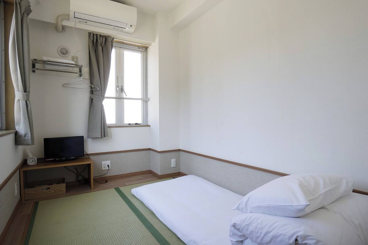 A hotel room with a single bed, a small desk with a television, an air conditioning unit, and a window with curtains.