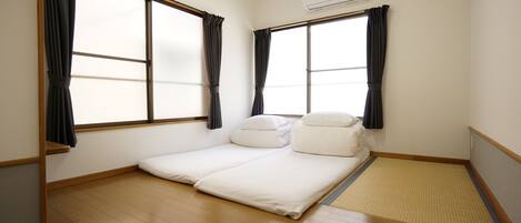Quarto com cama, duas janelas com cortinas, piso de madeira e ar-condicionado fixado na parede.