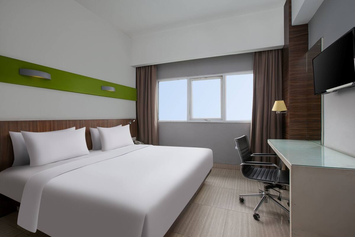 Quarto de hotel com uma cama grande, uma mesa com cadeira, uma TV de tela plana e uma janela com cortinas.