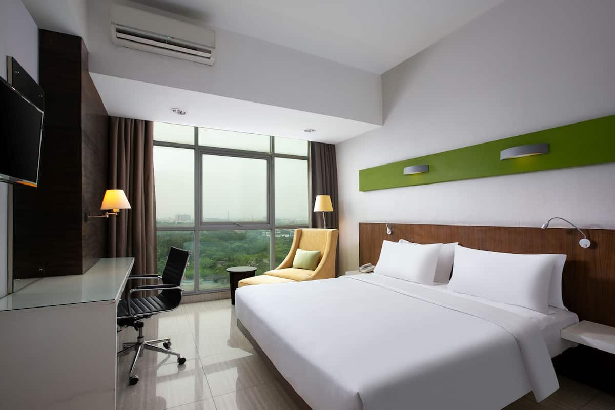 Quarto de hotel com uma cama grande, uma escrivaninha com cadeira, televisão e vista para a cidade pela janela.
