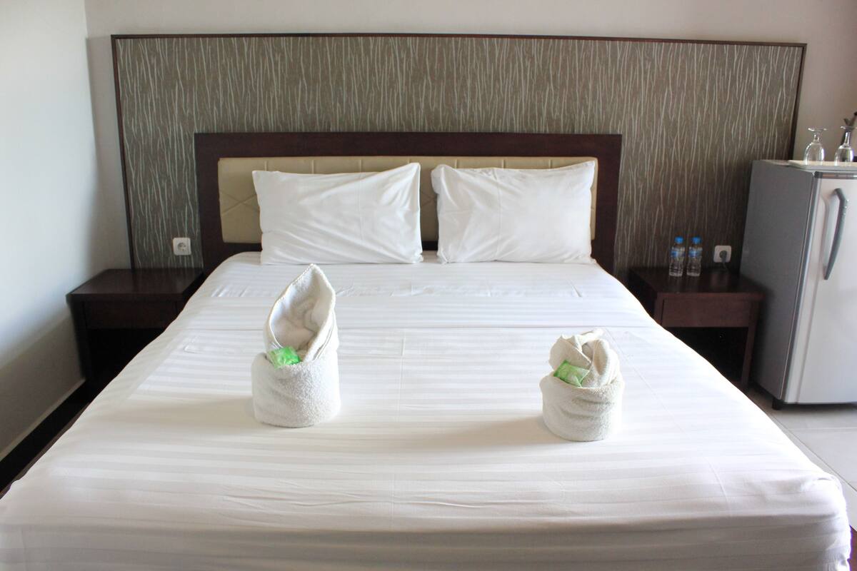 A neatly made bed with two towels placed on it, a nightstand with a lamp, and a refrigerator.