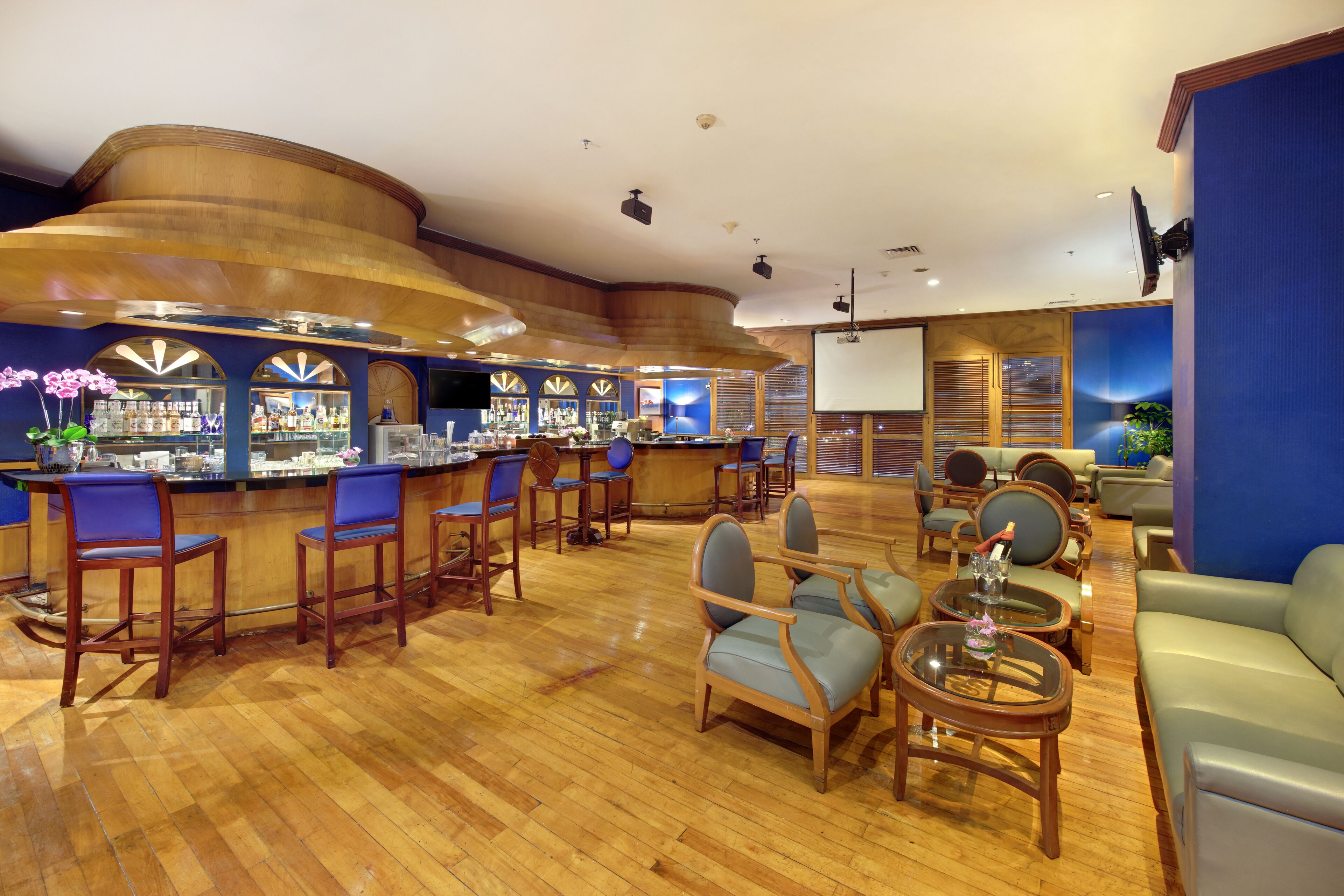 A bar area with wooden bar counters, blue cushioned chairs, and a seating area with leather sofas and round tables.