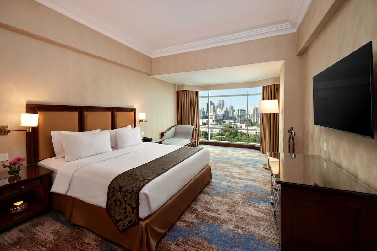 A hotel room with a large bed, a TV, a city view, and a desk with a lamp.